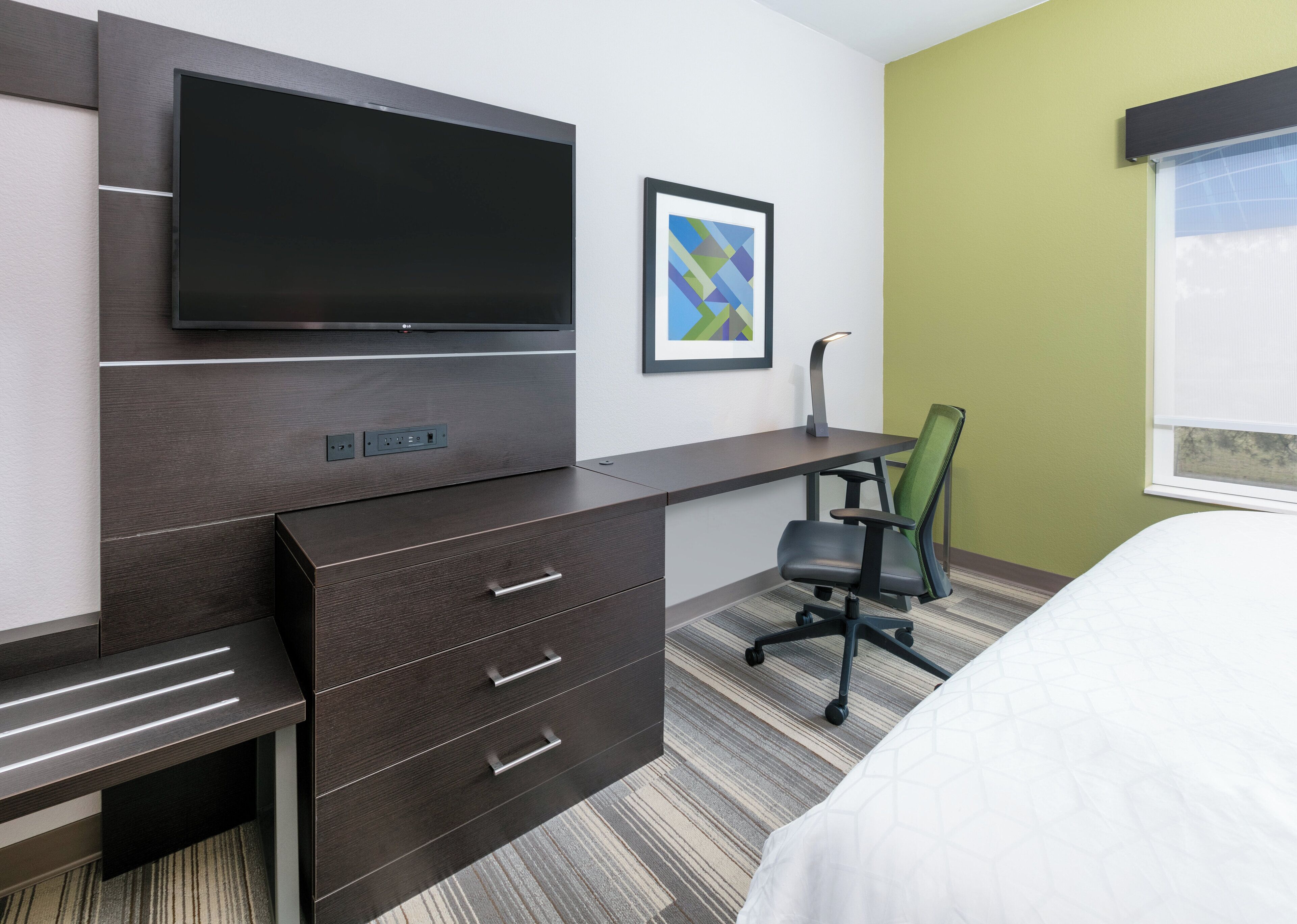 A hotel room with a dark wood desk, a flat-screen TV, a green chair, a bed, and a window with blinds.