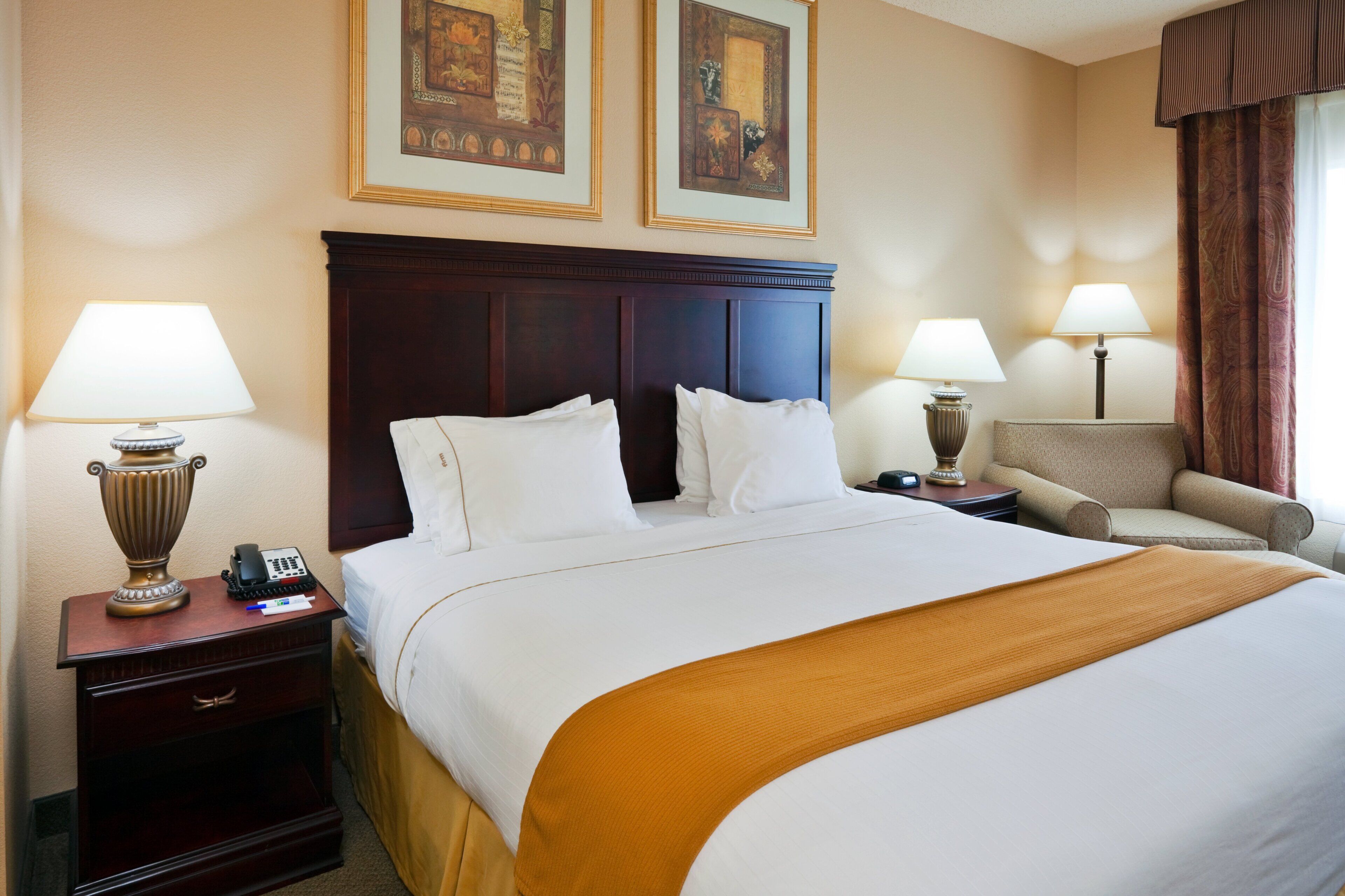 A hotel room with a large bed, two bedside lamps, a nightstand with a phone, a chair, and two framed pictures on the wall.