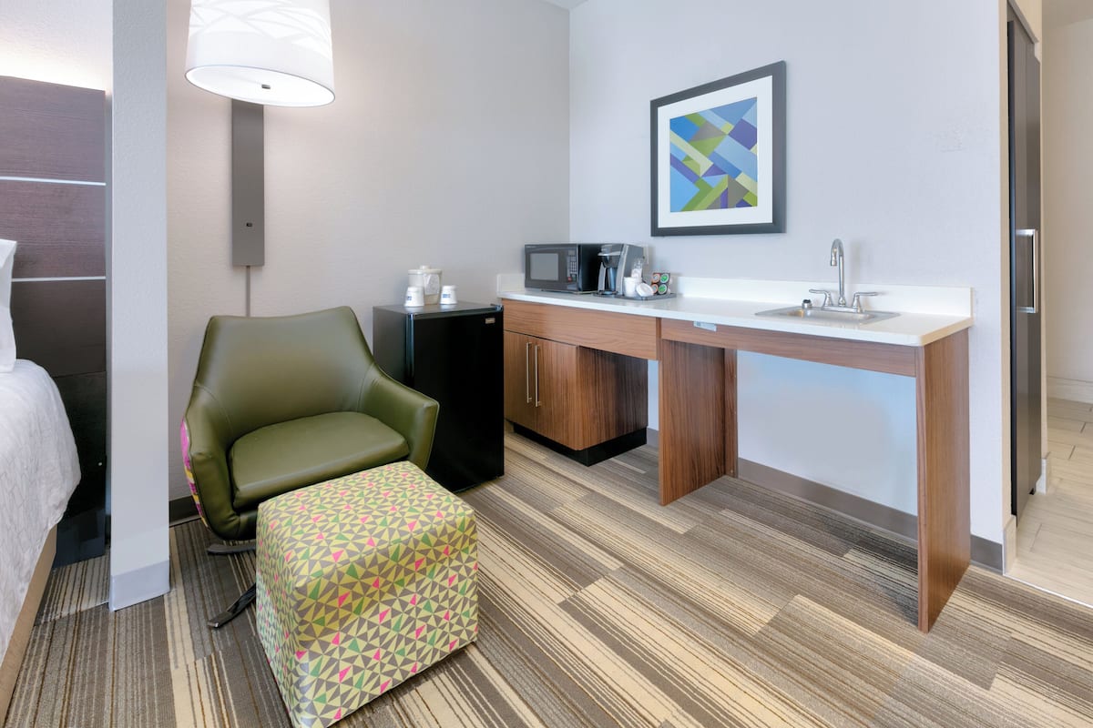 A hotel room with a bed, a green armchair, a small ottoman, a desk with a microwave, a coffee maker, and a sink.