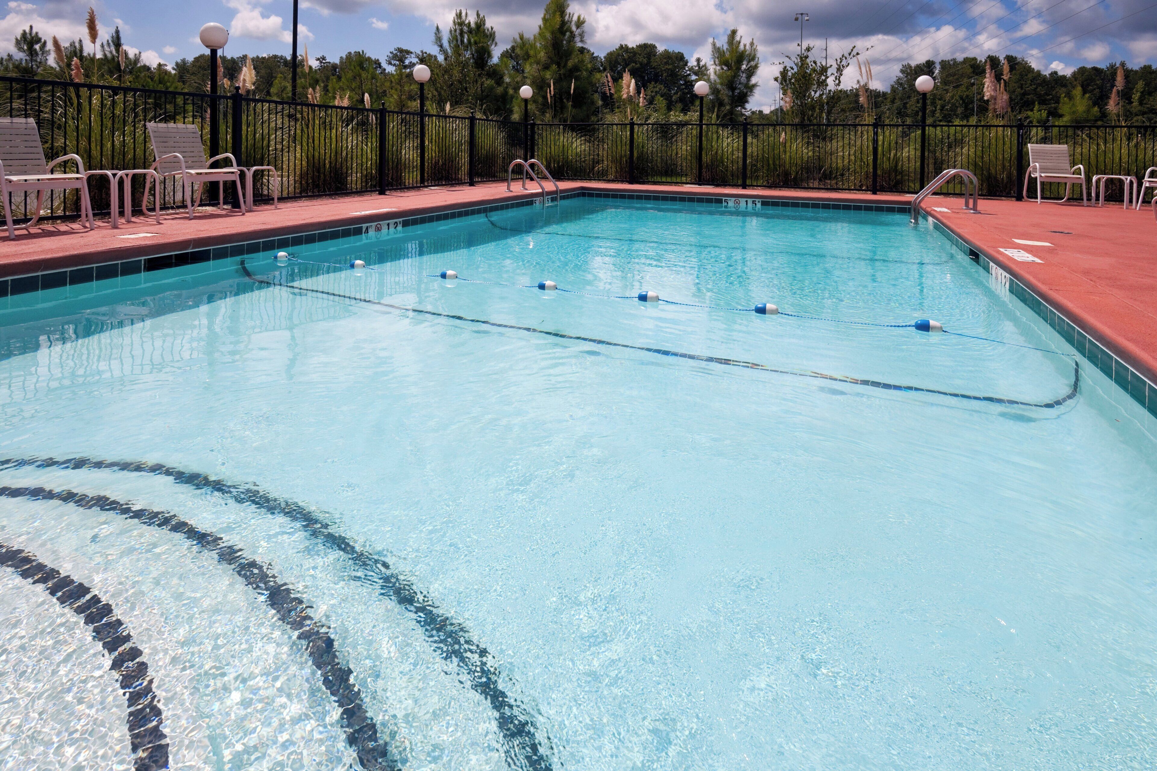 A clear swimming pool with lane dividers, surrounded by a black fence and equipped with lounge chairs.