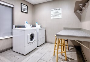 A guest laundry area with a washing machine, dryer, and a table with a stool. A sign above the machines states 'Guest Laundry' with usage instructions. - Holiday Inn Express Hotel & Suites Pell City by IHG (Pell City)