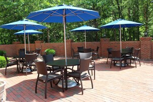 An outdoor patio with brick flooring, wicker chairs, and tables under blue umbrellas.