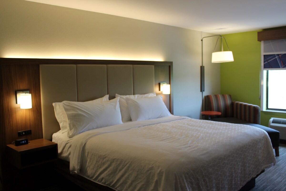 A hotel room with a large bed, a nightstand with a clock, a chair, and a lamp.