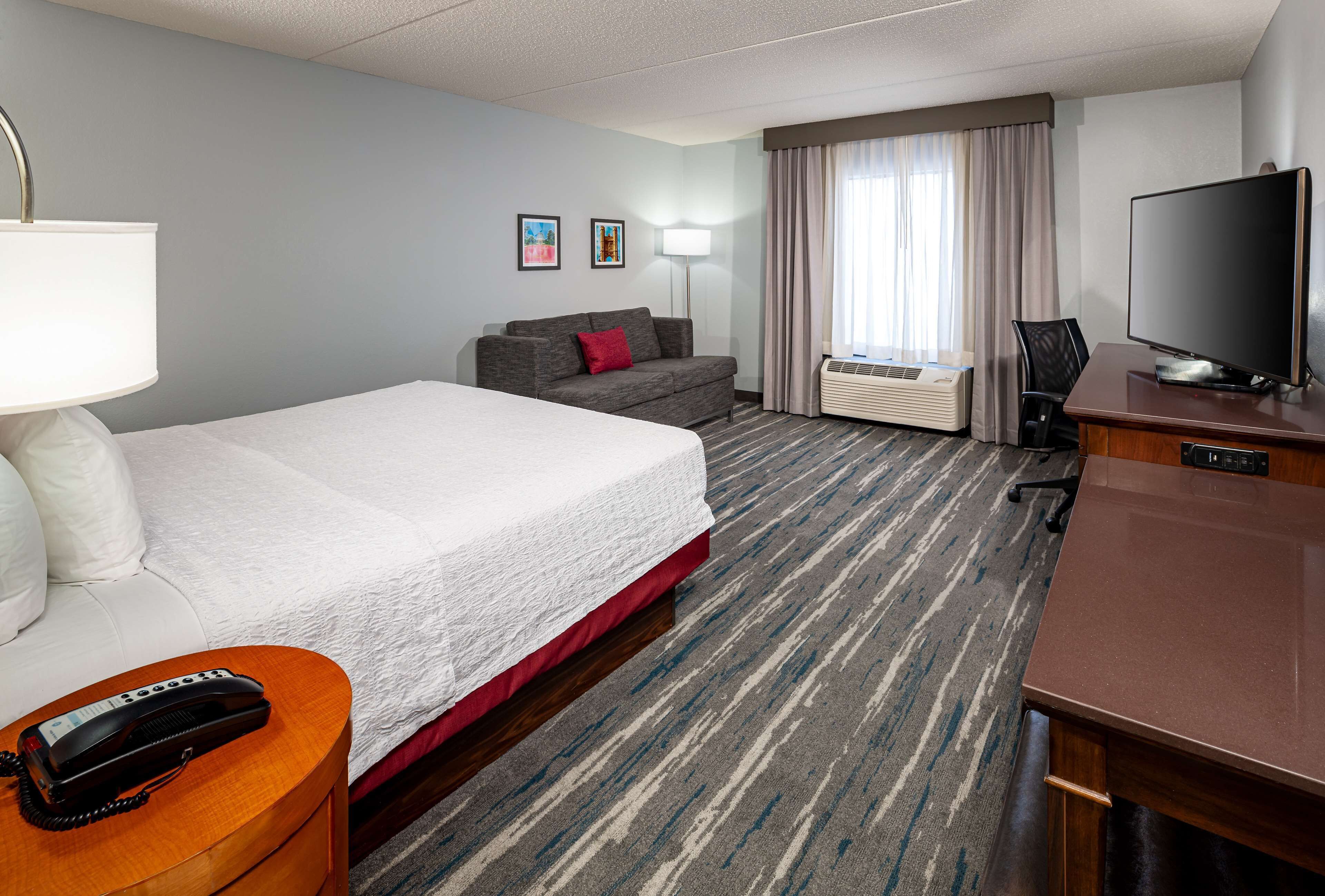 A hotel room with a large bed, a sofa, a desk with a TV, a lamp, and an air conditioning unit.