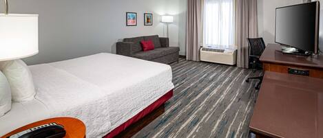 A hotel room with a large bed, a sofa, a desk with a TV, a lamp, and an air conditioning unit.