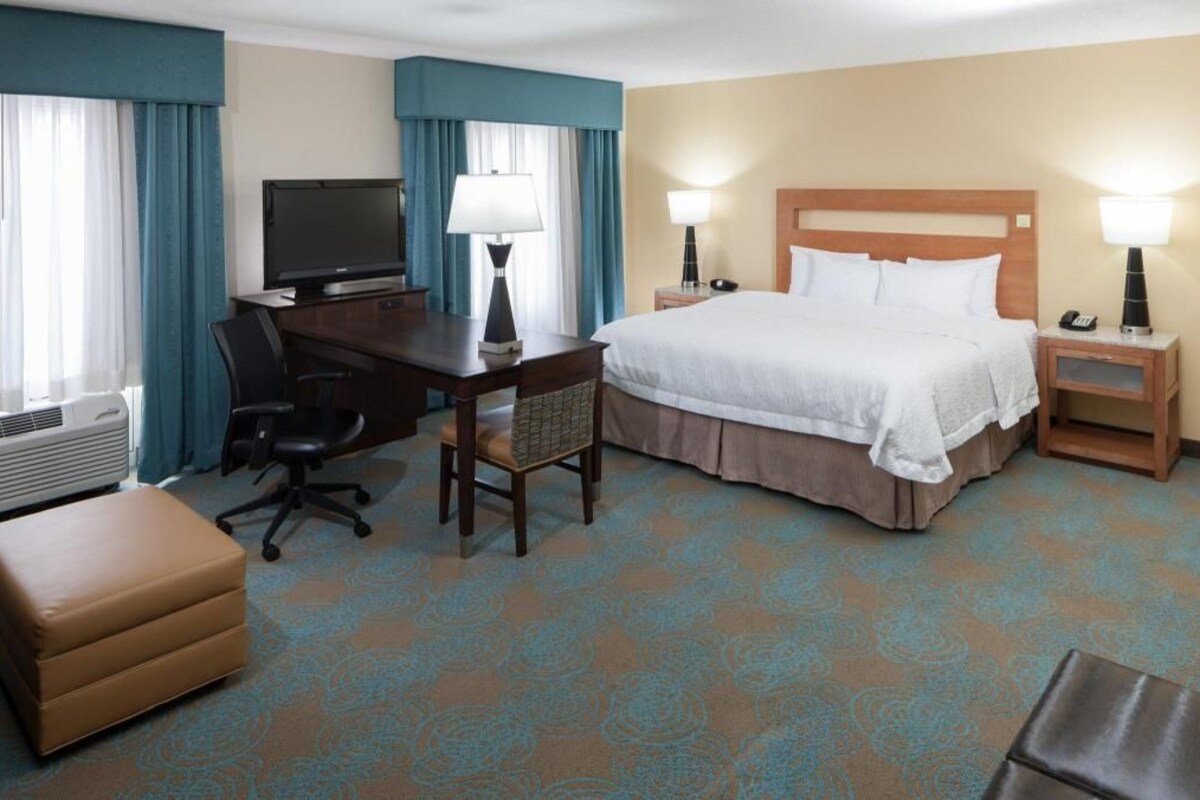 Habitación de hotel con cama, escritorio, silla, televisor, lámparas y una pequeña mesa.