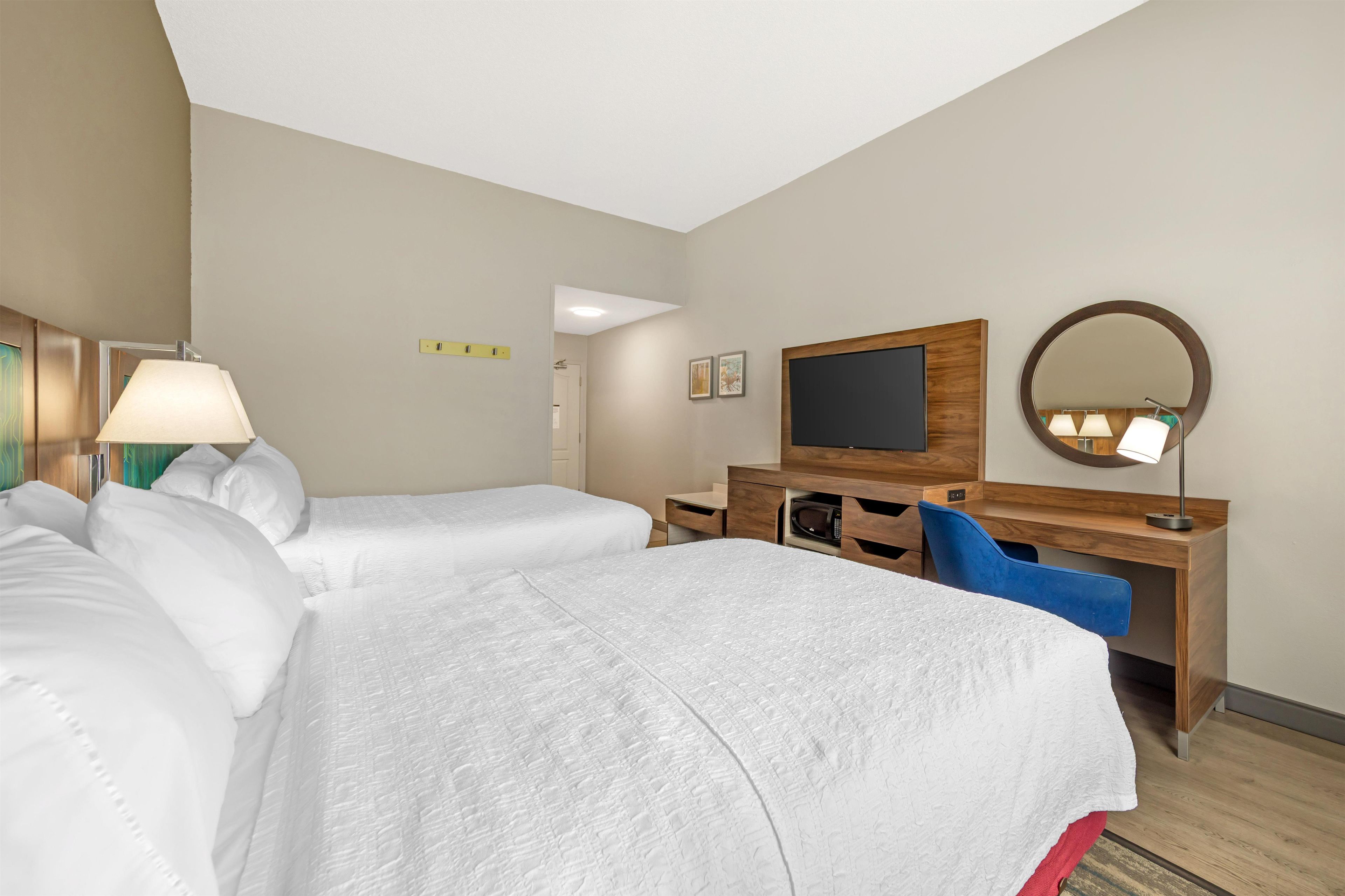 Habitación de hotel con dos camas, un escritorio con televisor, un espejo y una lámpara.