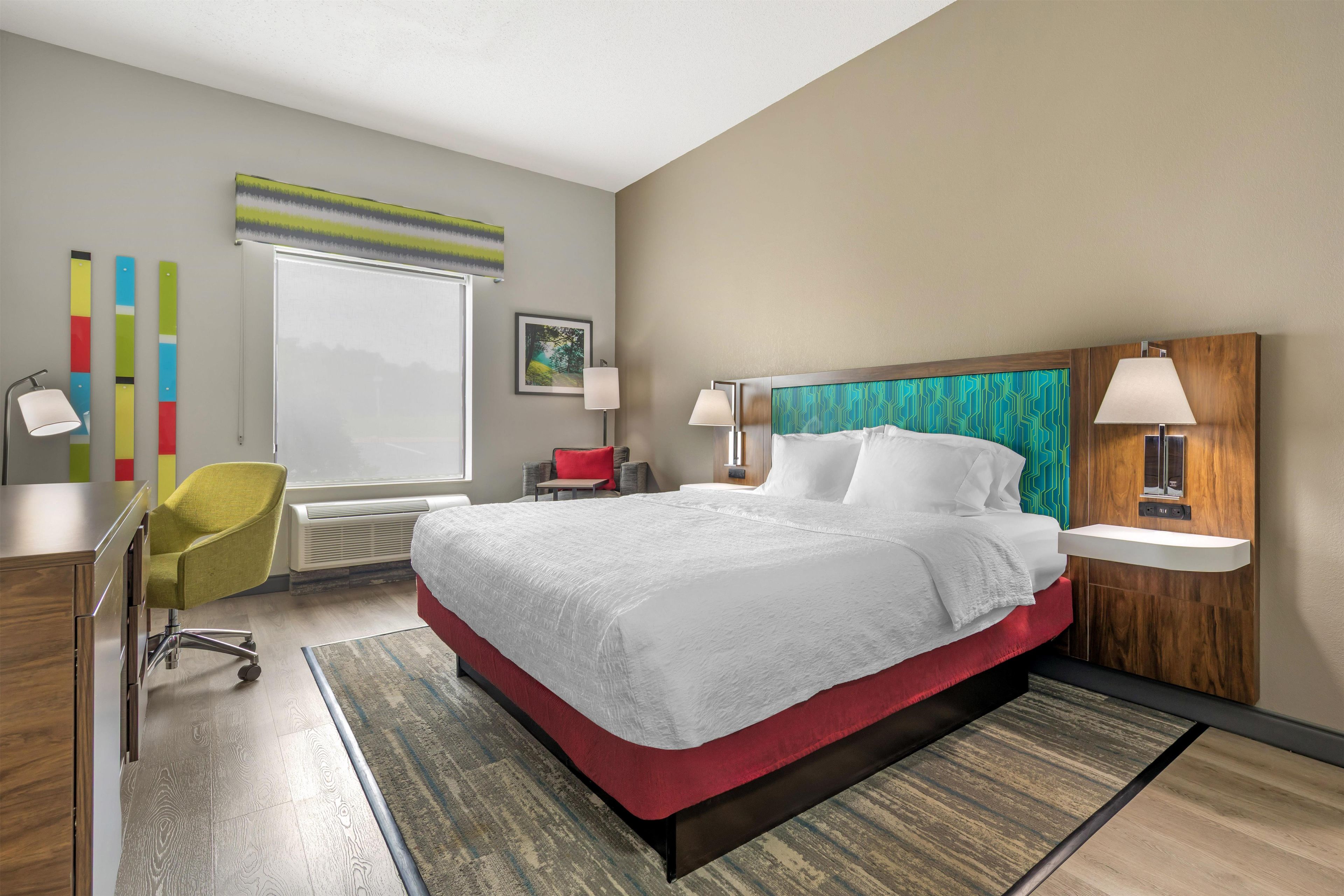 A hotel room with a bed, a desk, a chair, a window, and a wall with a picture.