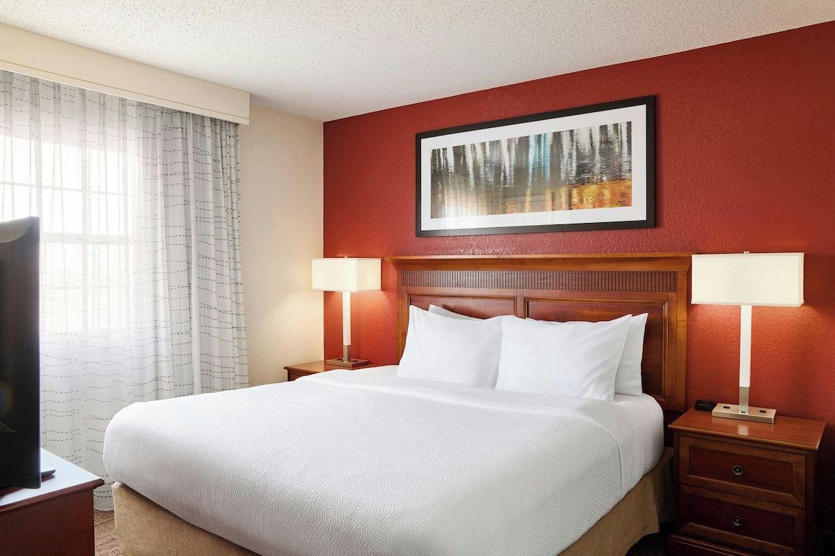 A hotel room with a large bed, two bedside tables with lamps, a television on the left side, and a framed picture on the wall.