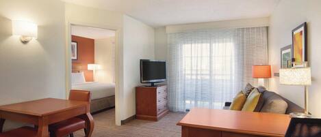 A hotel room with a wooden desk, a sofa, a television on a wooden cabinet, and a bed with a white headboard.