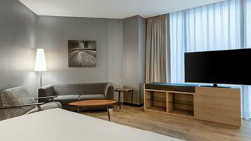 Una habitación de hotel moderna con sofá, una mesita redonda, un televisor sobre un mueble de madera y un cuadro en la pared.
