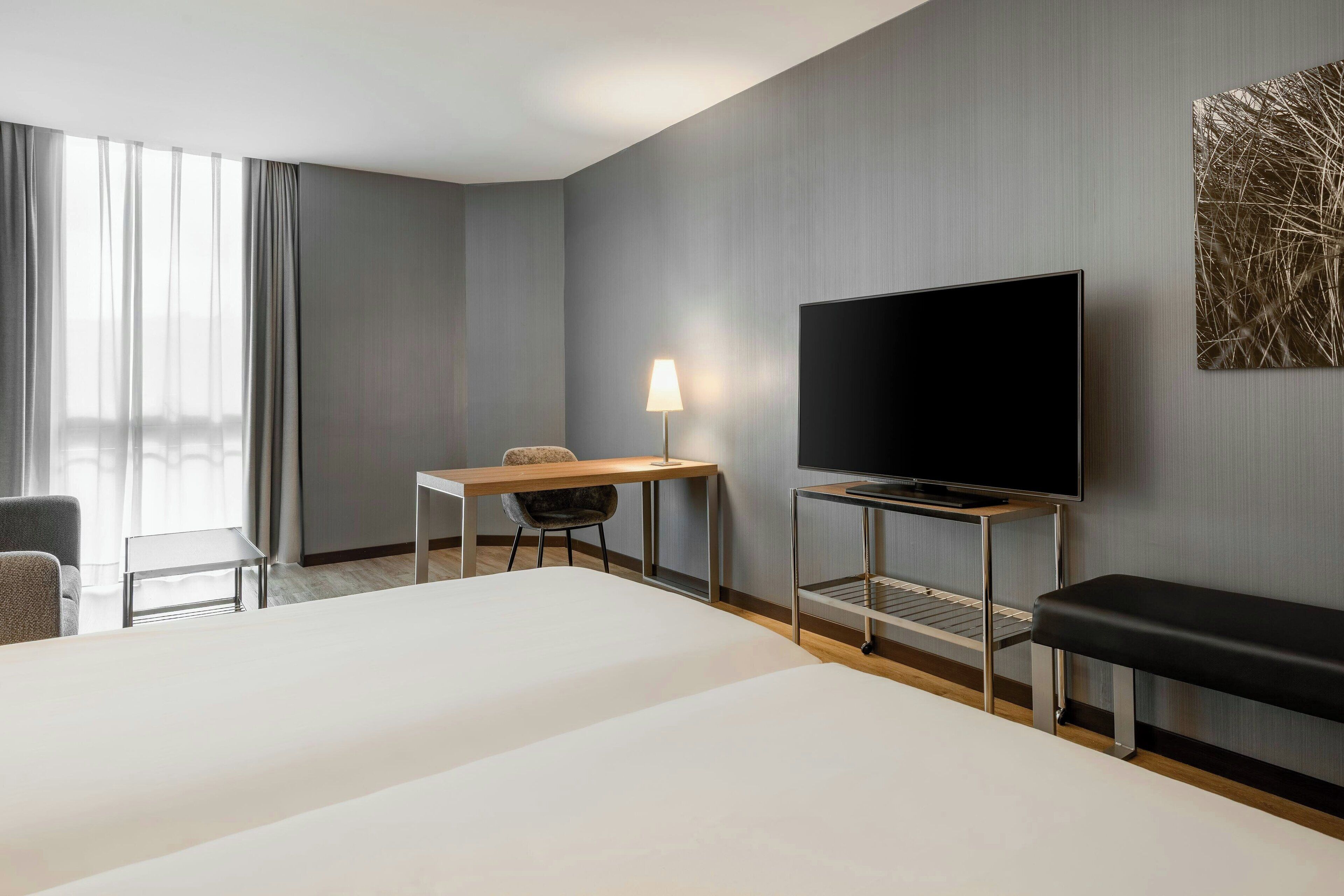 Quarto de hotel com uma cama grande, uma escrivaninha com cadeira, uma televisão e um banco.