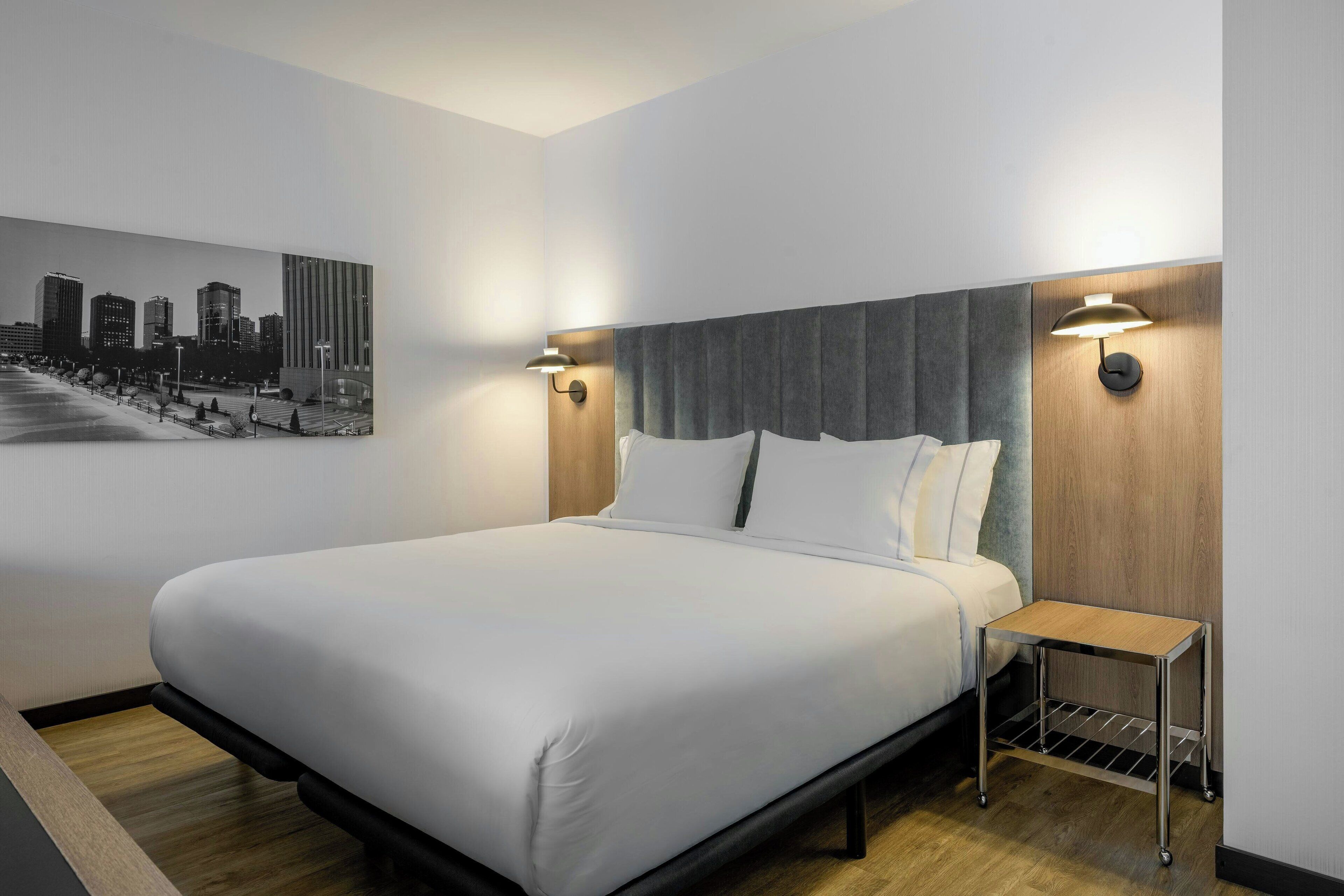 Una habitación de hotel moderna con una cama grande, una mesita de noche y un cuadro de un paisaje urbano en la pared.