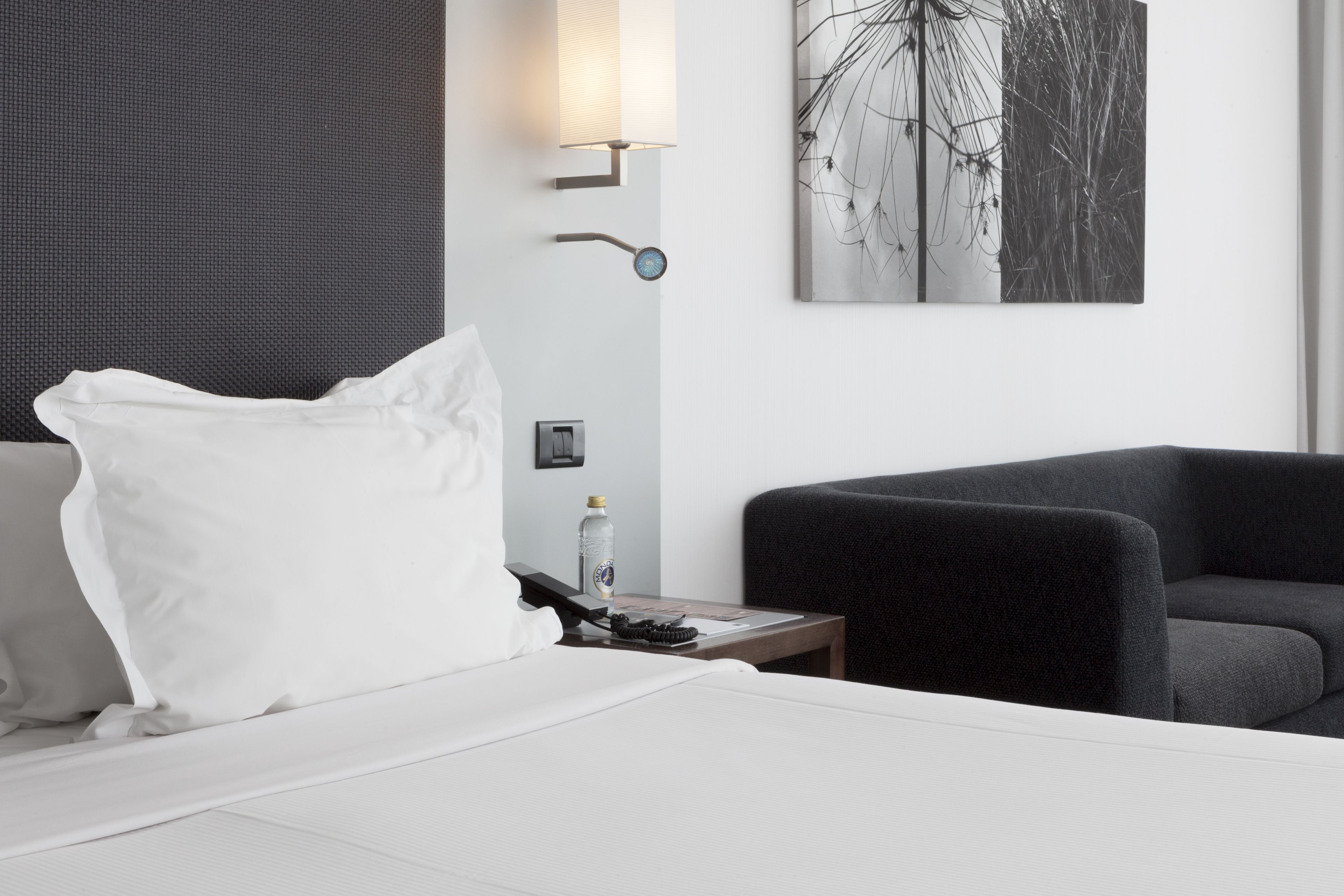 A hotel room with a bed, a nightstand, a water bottle, a remote control, and a wall-mounted lamp.