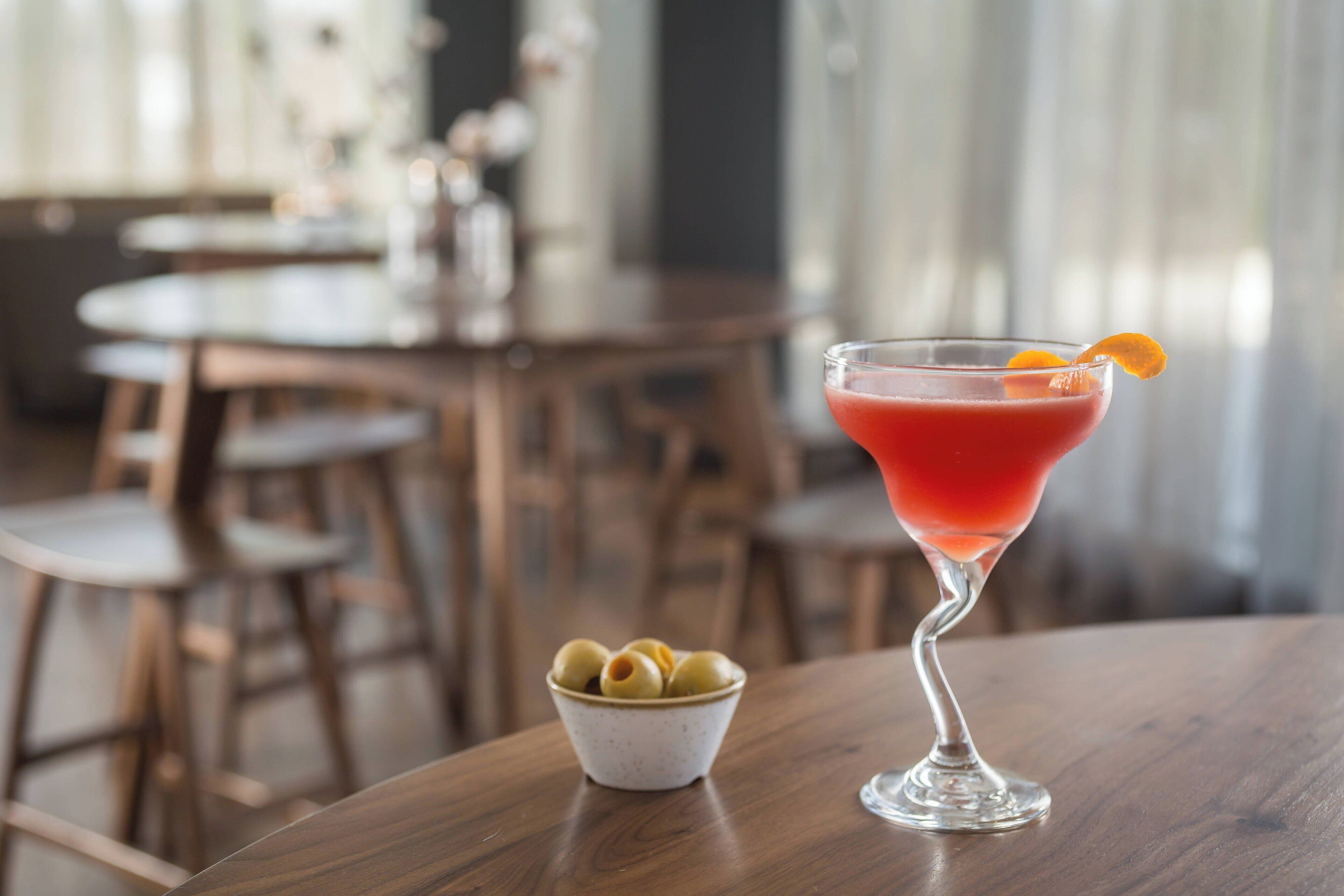 Una copa de martini con una bebida roja y una cáscara de naranja sobre una mesa de madera.