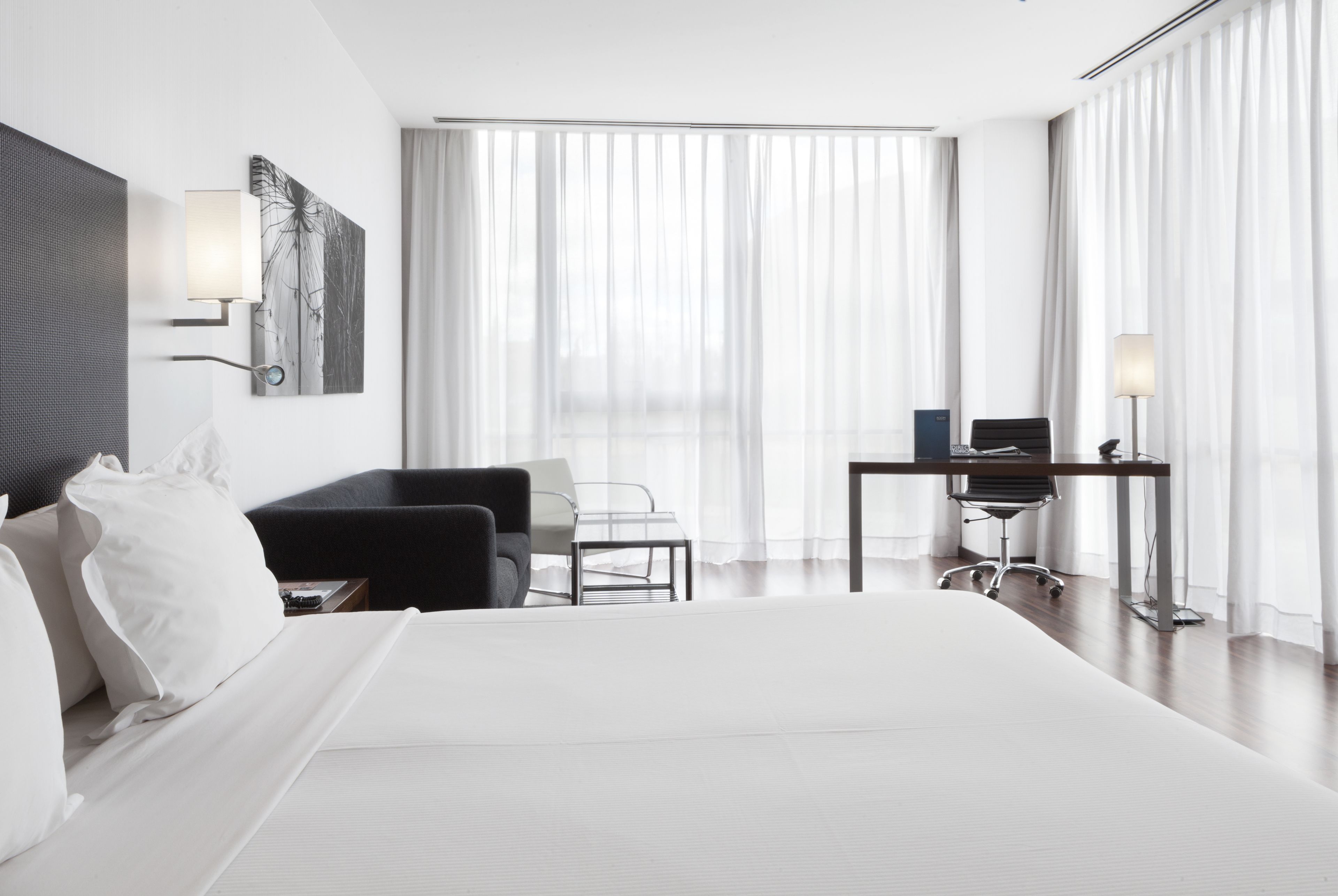 A modern hotel room with a large bed, a desk with a chair, a sofa, and a wall-mounted lamp.