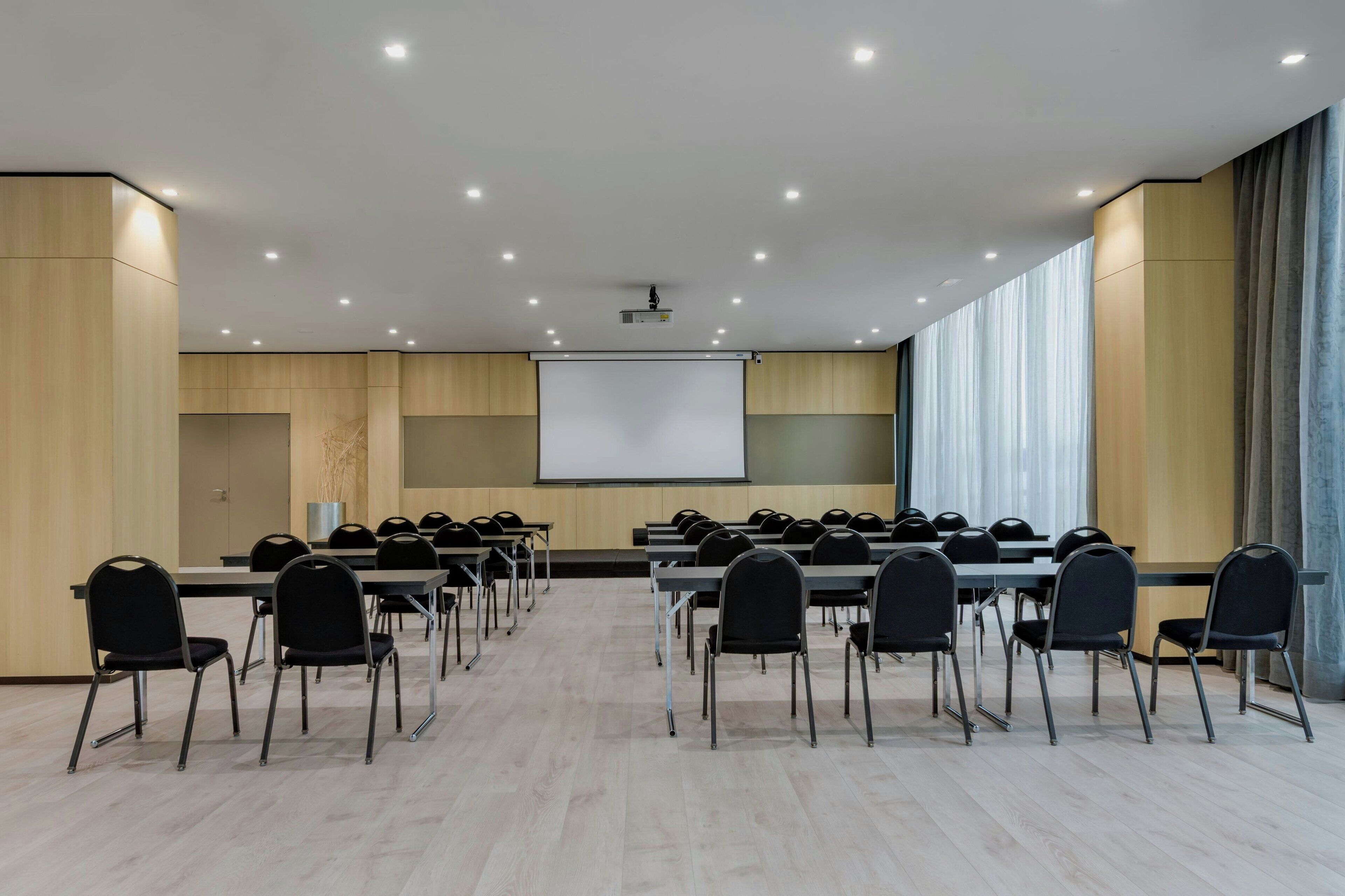 Ein Konferenzraum mit mehreren Tischen und Stühlen, einer Projektionsleinwand und einem großen Fenster mit Vorhängen.