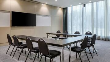 Une salle de conférence avec une longue table, des chaises, un téléviseur à écran plat et une grande fenêtre avec des rideaux.