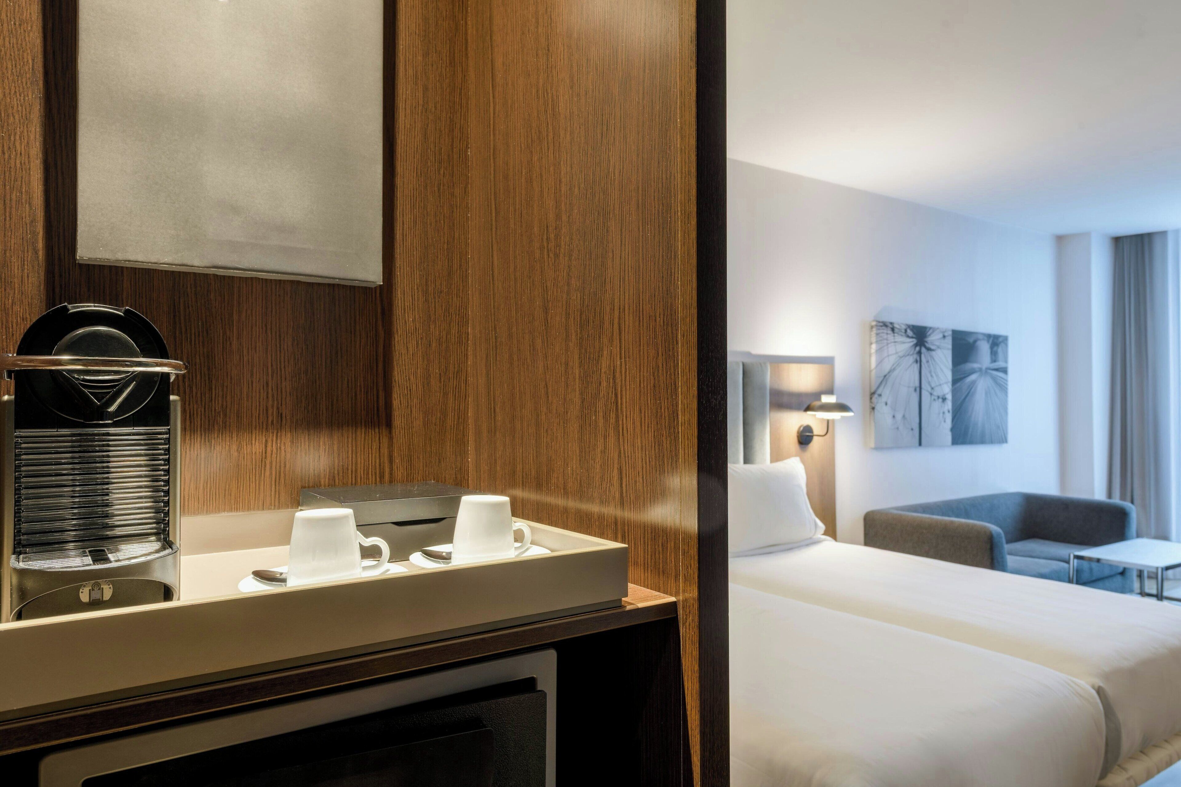 Habitación de hotel con pared de paneles de madera, una cafetera, una bandeja con tazas, una cama, un sofá y una lámpara.