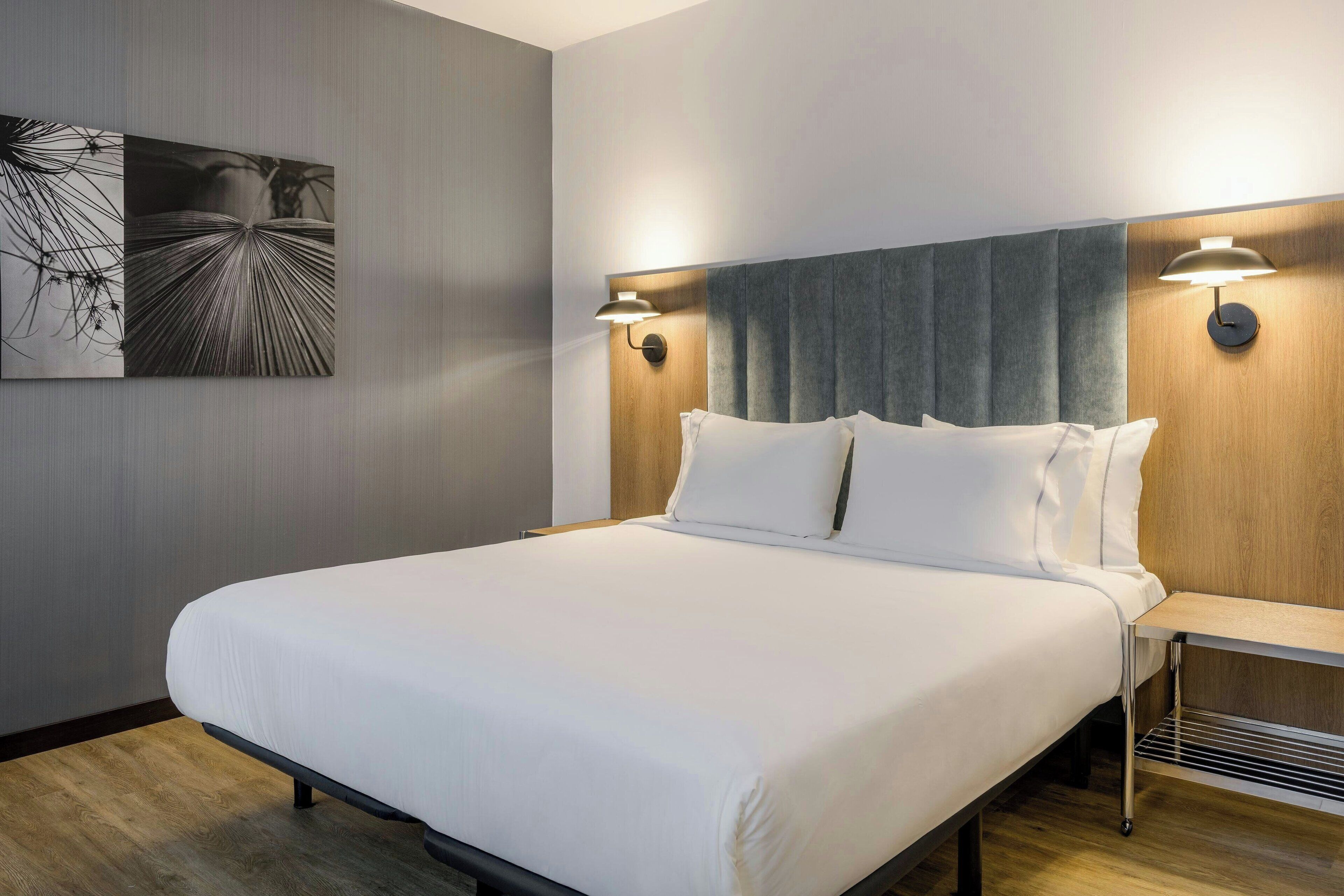 Una cama bien hecha con sábanas blancas, cabecera con panel gris y dos lámparas fijadas a la pared a cada lado.