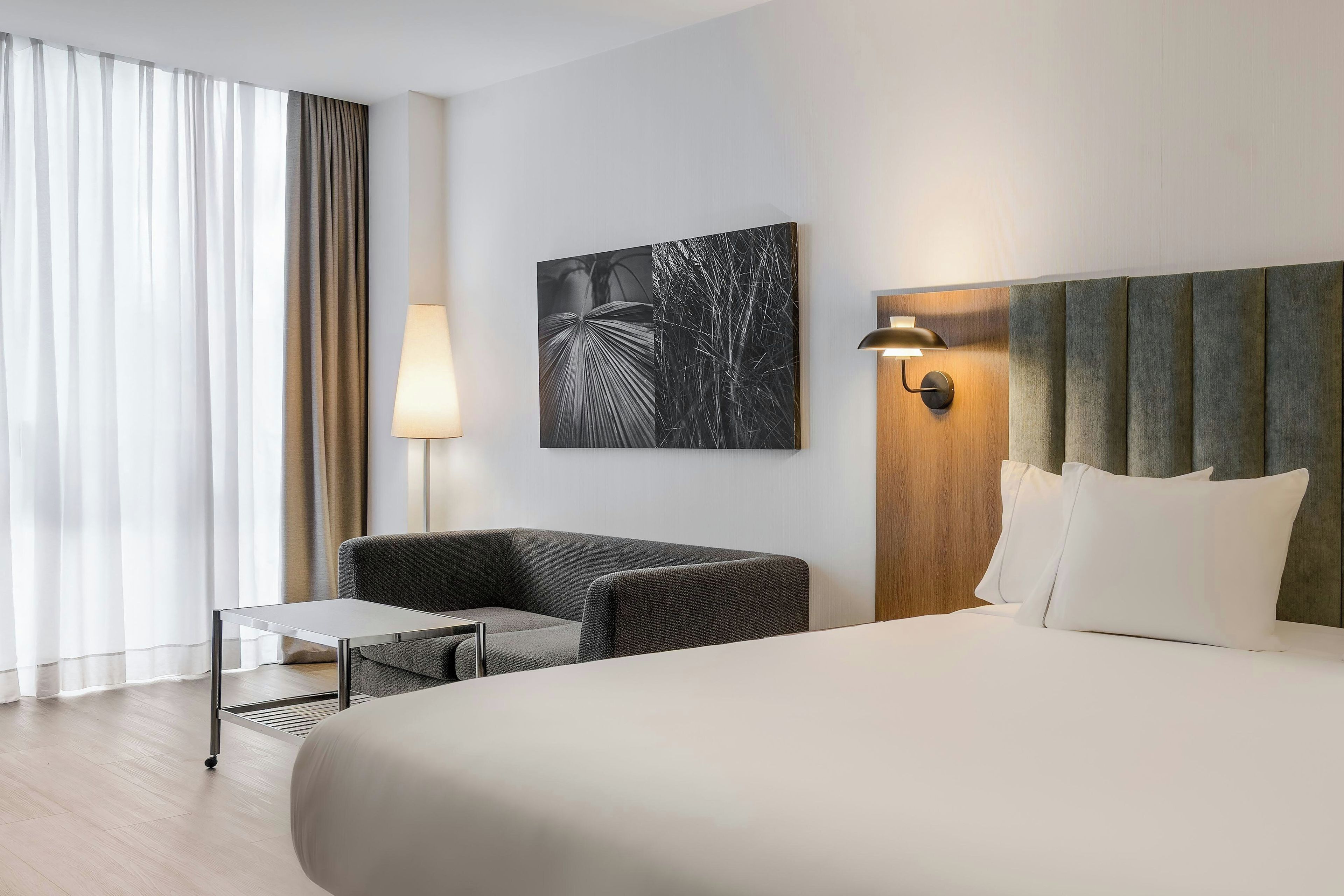 Habitación de hotel con cama, una silla, una mesita, una lámpara y una obra de arte colgada en la pared.