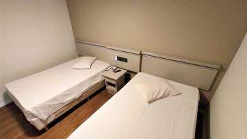 A small, clean room with two single beds, a bedside table with a phone, and a wall-mounted light switch.
