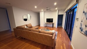 Una sala de estar moderna con un sofá grande en forma de L, un televisor de pantalla plana sobre un mueble, piso de madera y un ventilador de techo.