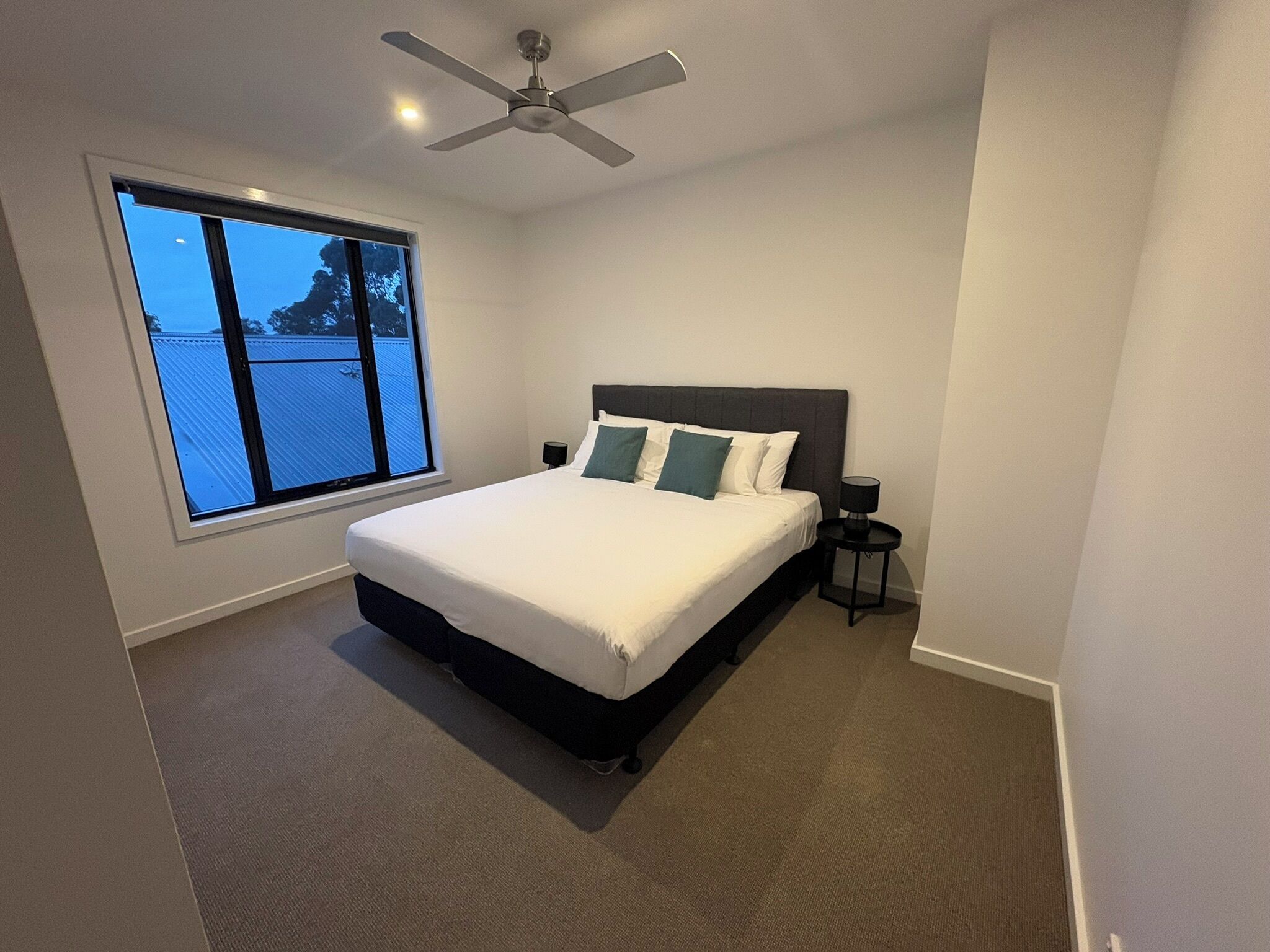 A bedroom with a large bed, a ceiling fan, a window with a view, and a nightstand with a lamp.
