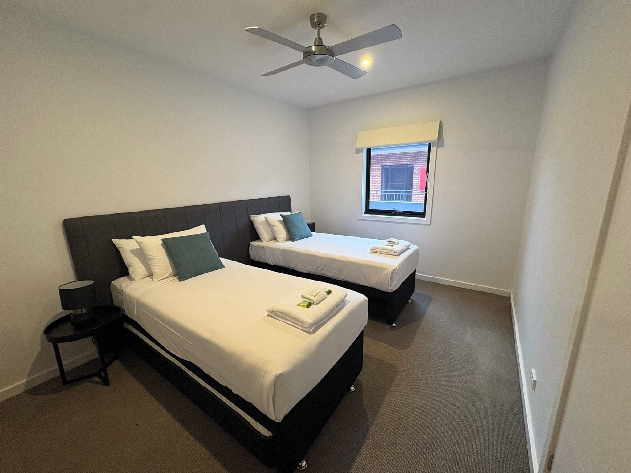 Habitación de hotel con dos camas, un ventilador de techo, una mesita de noche y ventana con vista a un edificio.