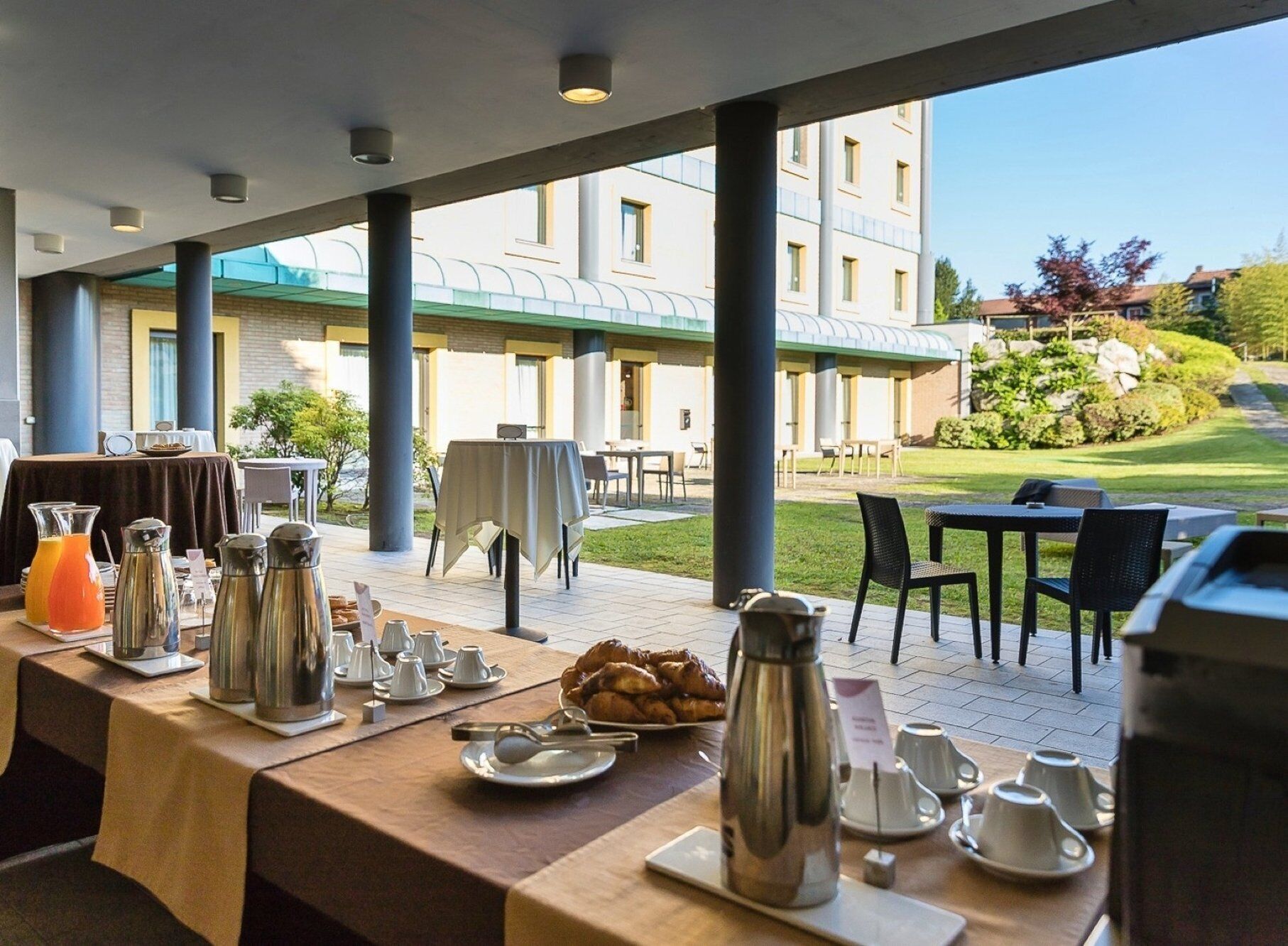 A breakfast setup with tables, coffee pots, and a view of a building and garden.