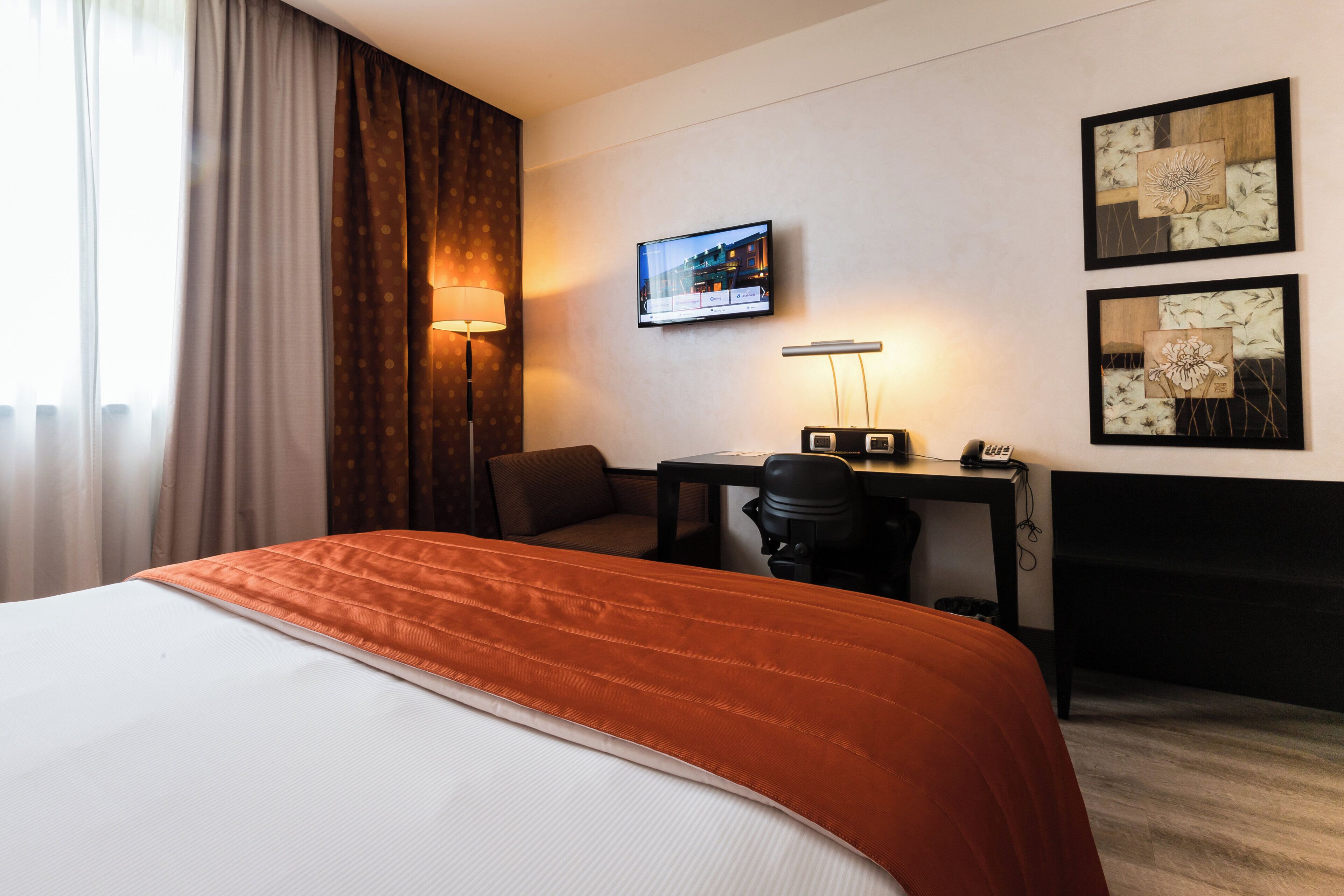 A hotel room with a bed, a desk, a chair, a TV, and artwork on the wall.
