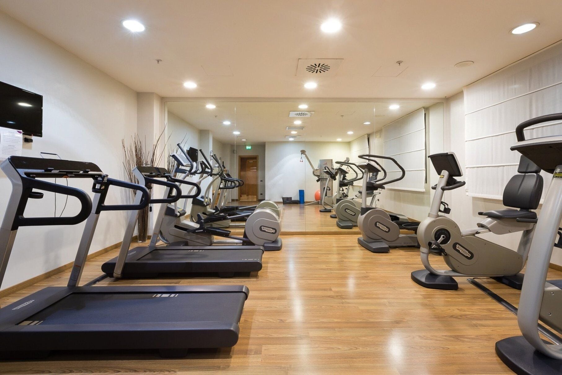 Un gimnasio con varias caminadoras y elípticas, suelo de madera y un espejo grande.