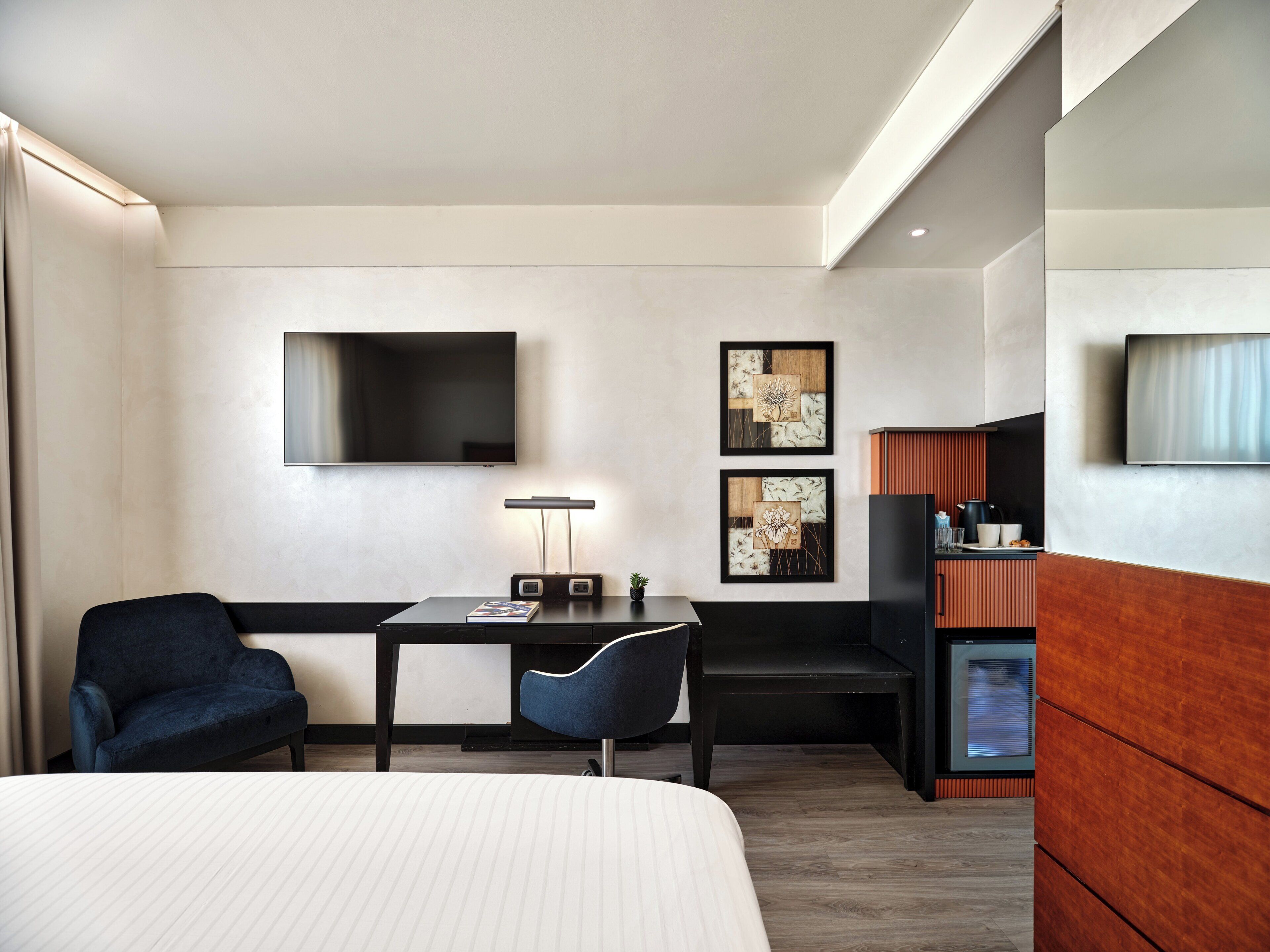 A modern hotel room with a bed, a desk, a chair, a TV, and artwork on the wall.