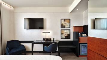 A modern hotel room with a bed, a desk, a chair, a TV, and artwork on the wall.