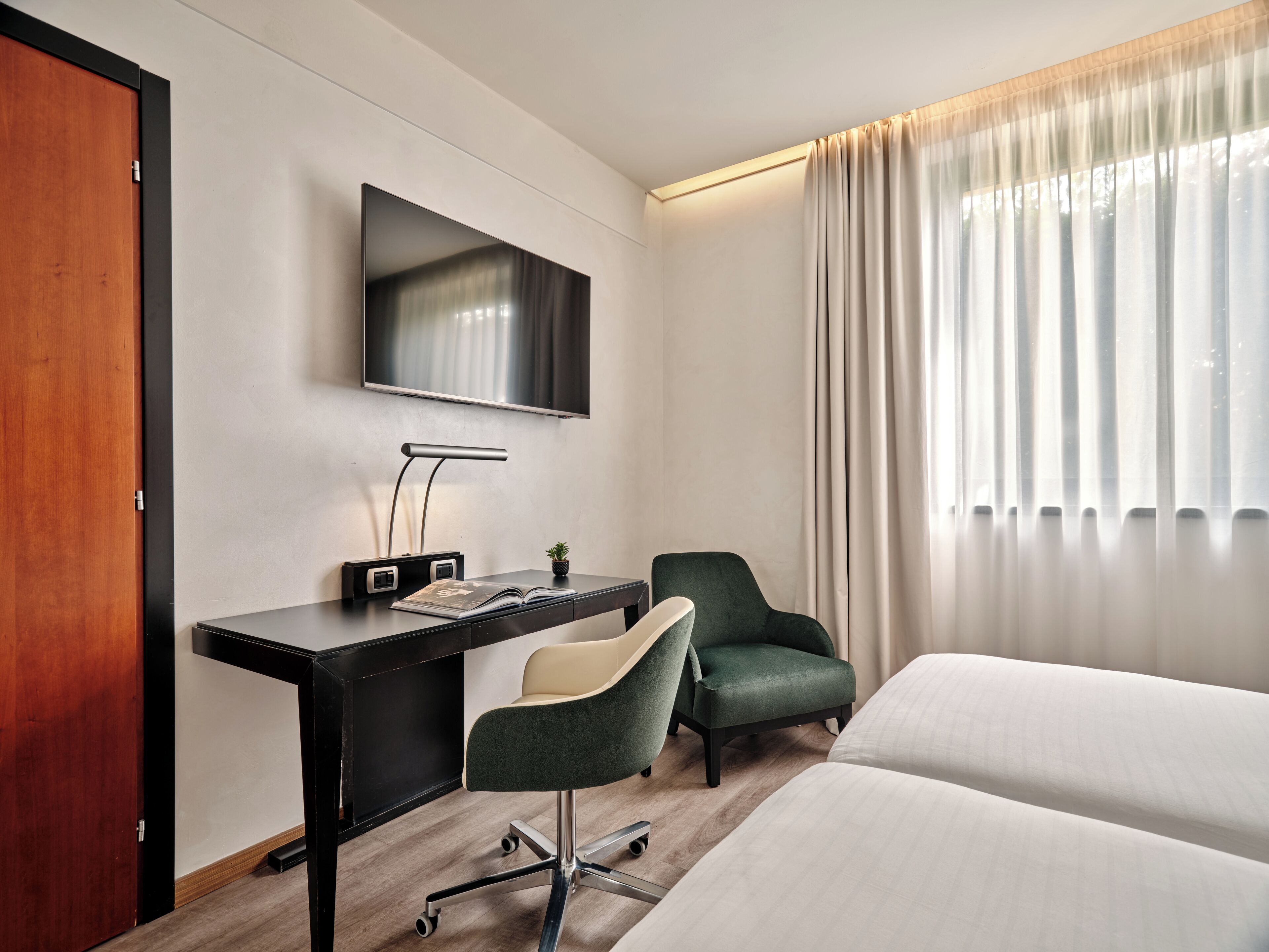 A hotel room with a bed, a desk with a chair, a television, and a window with curtains.