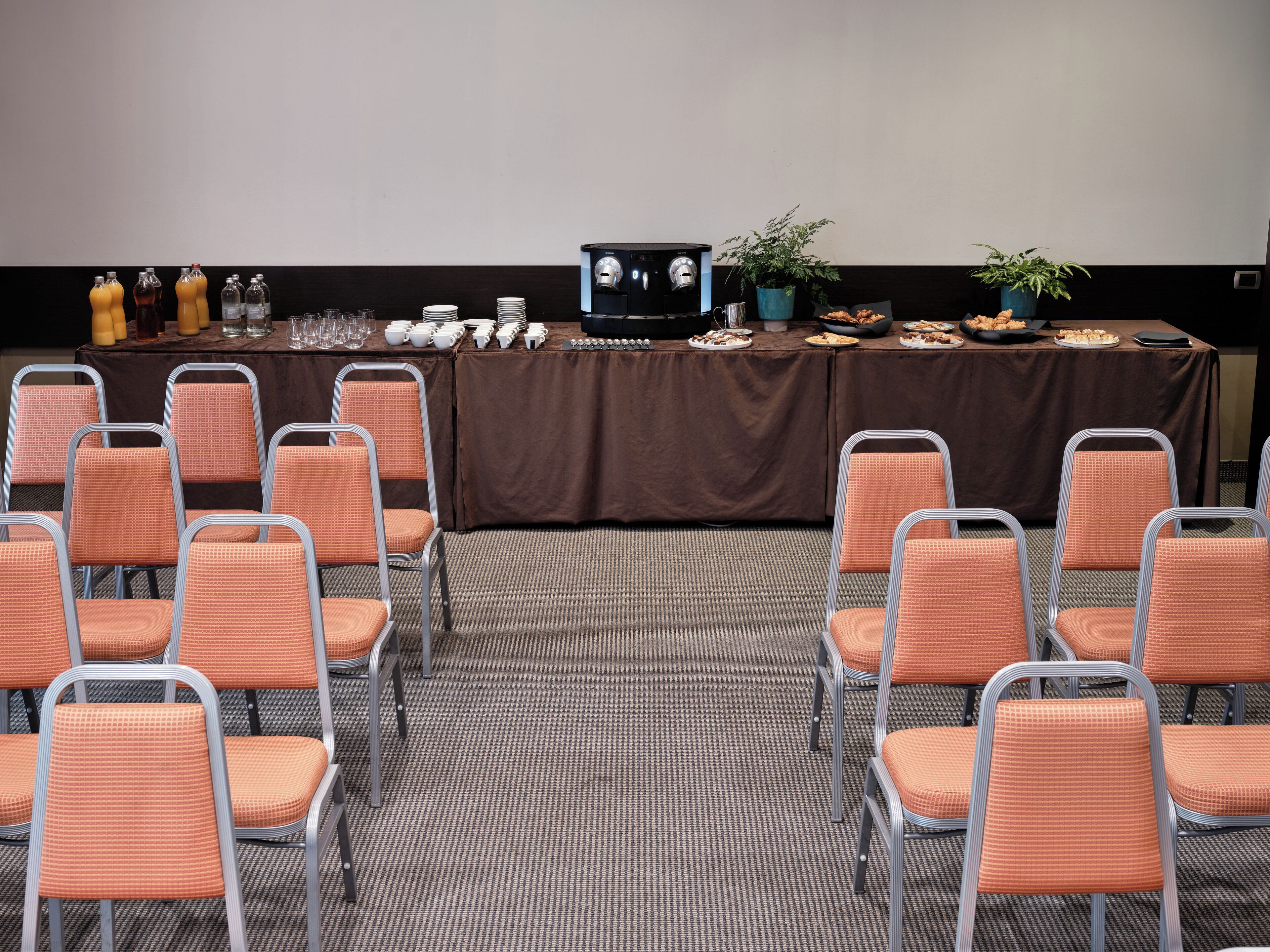 Un salón de conferencias con filas de sillas anaranjadas, una mesa marrón con diversos objetos y una pantalla de proyector.