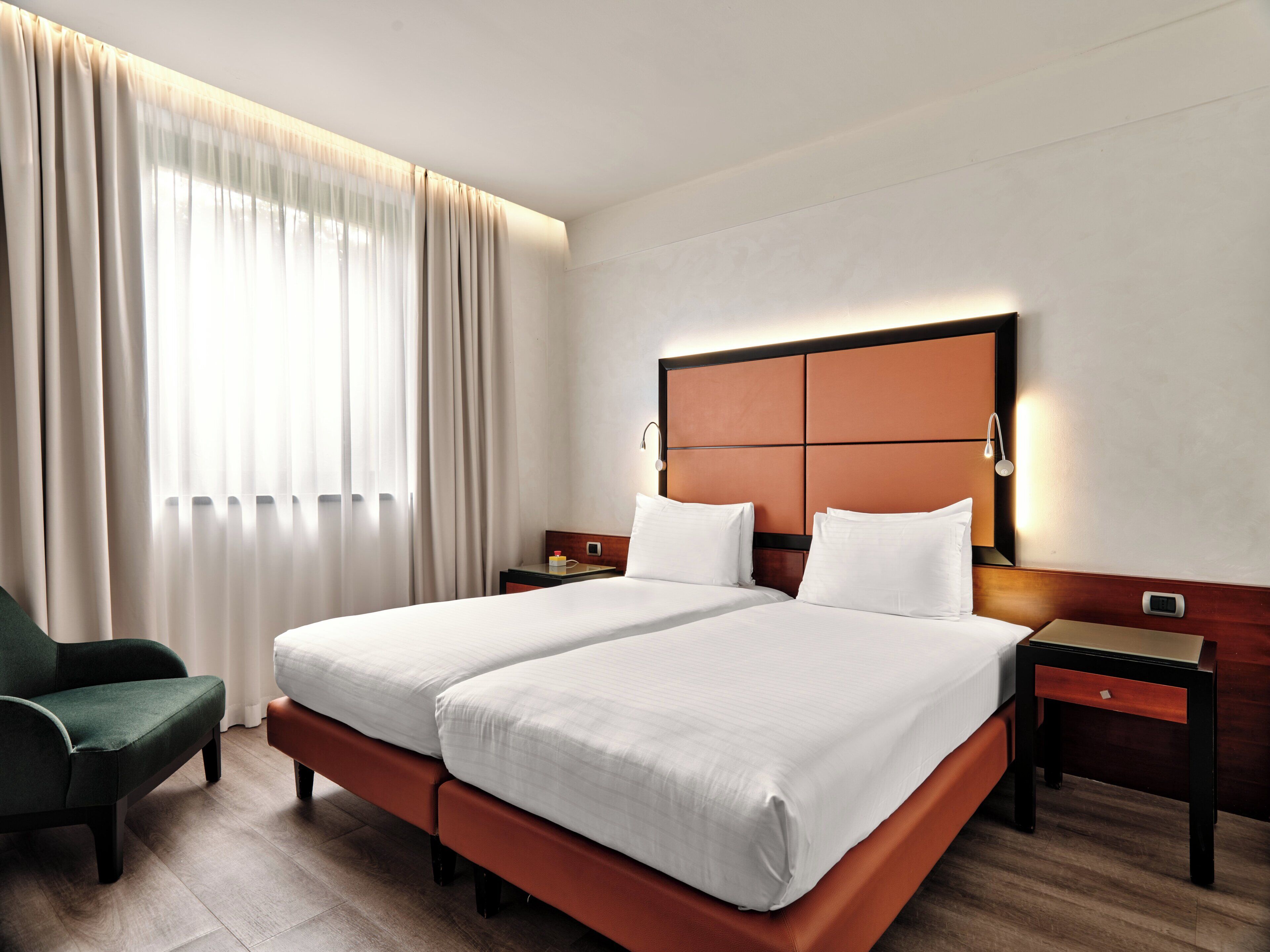 Habitación de hotel con cama doble, mesita de noche, una silla y un ventanal con cortinas.
