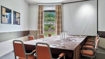 A conference room with a long table, chairs, a projector screen, and a view of greenery outside.