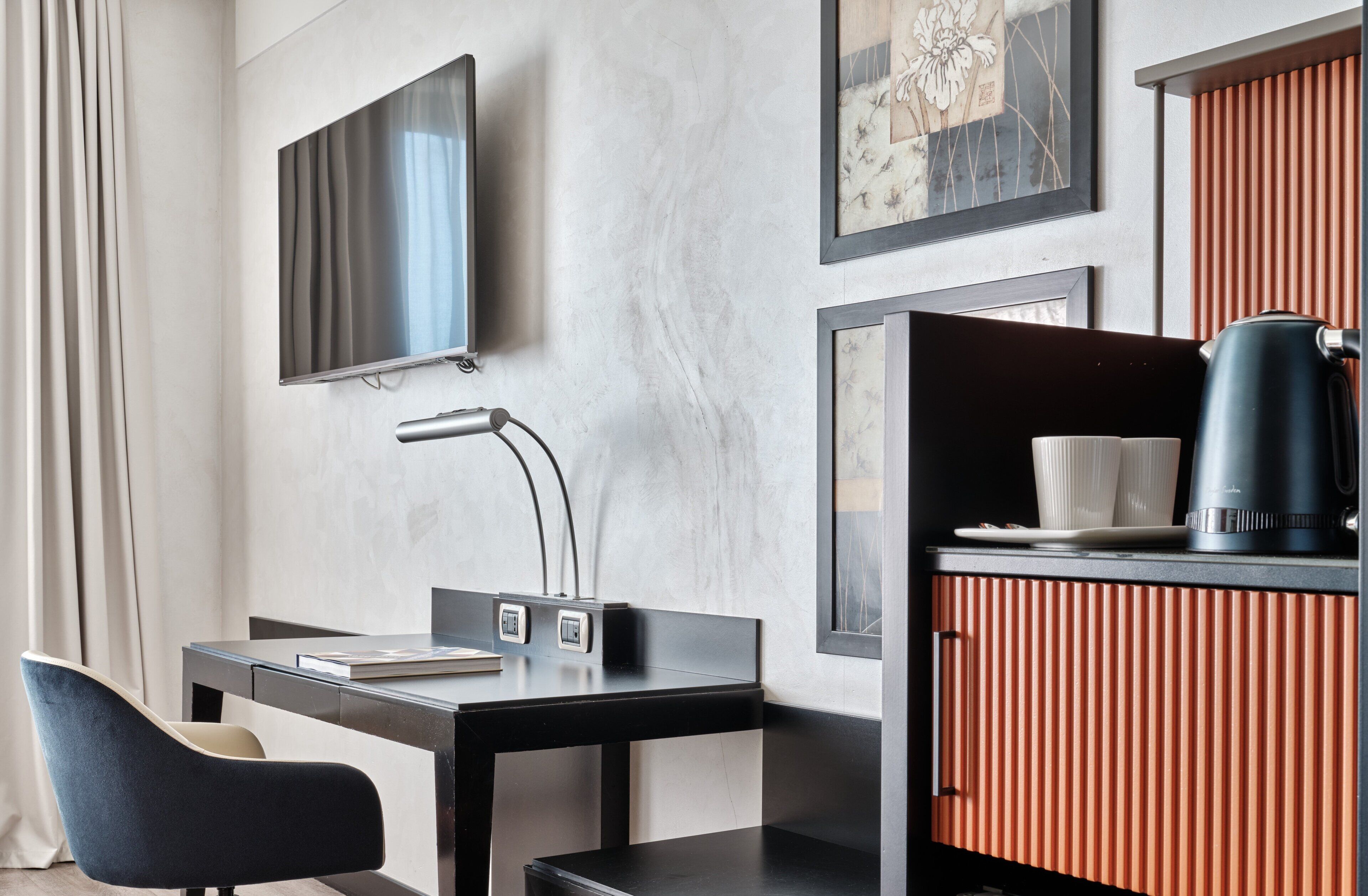 Habitación de hotel moderna con una televisión de pantalla plana, un escritorio con lámpara, una silla, cafetera y una obra de arte enmarcada en la pared.