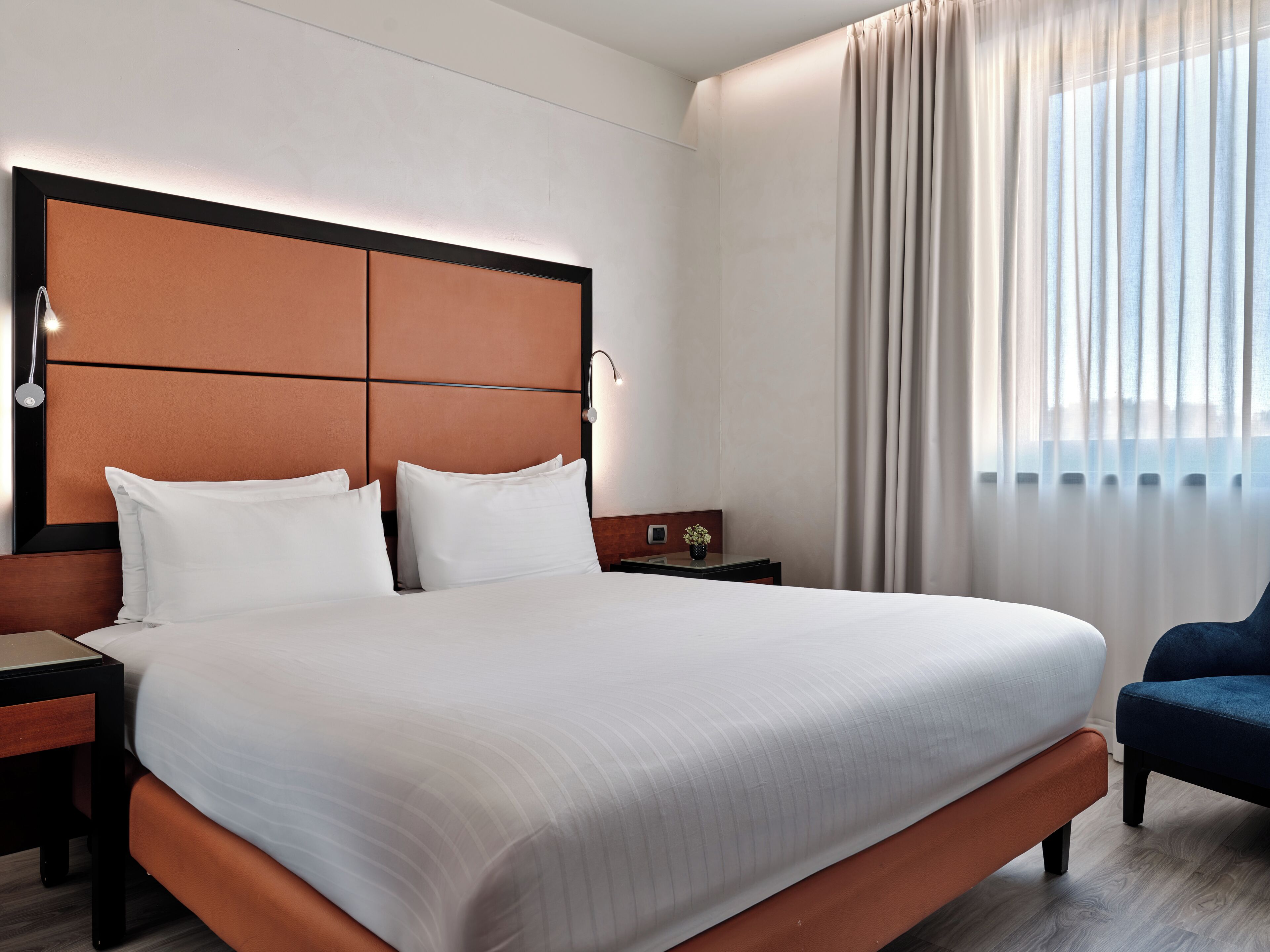 Habitación de hotel moderna con una cama grande, una mesita de noche, una lámpara y vistas de la ciudad a través de amplios ventanales.