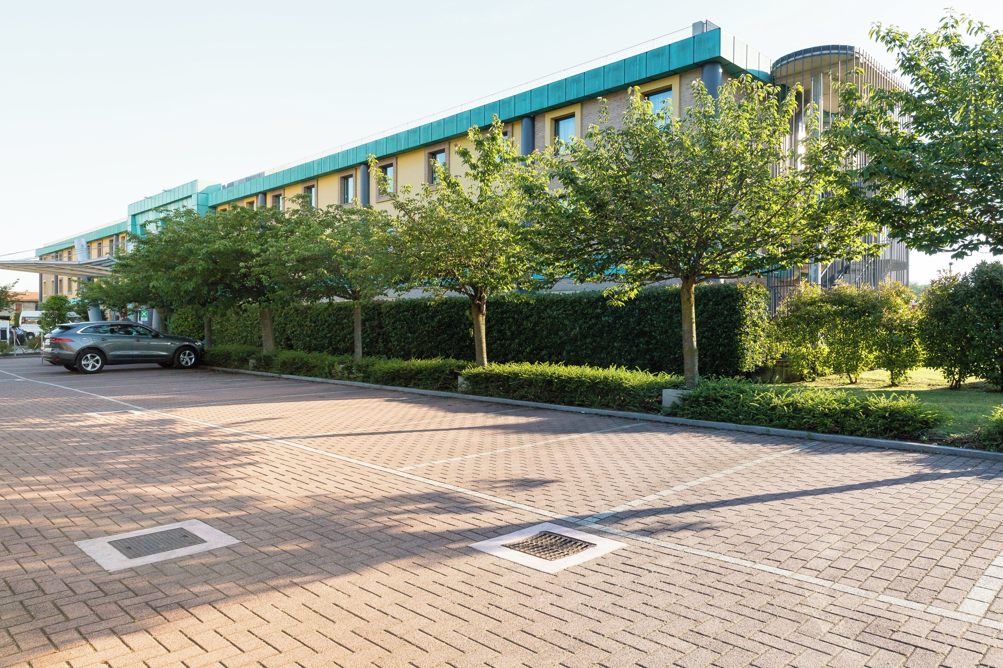 A paved parking area with a car parked, a building with a blue roof, and green trees.