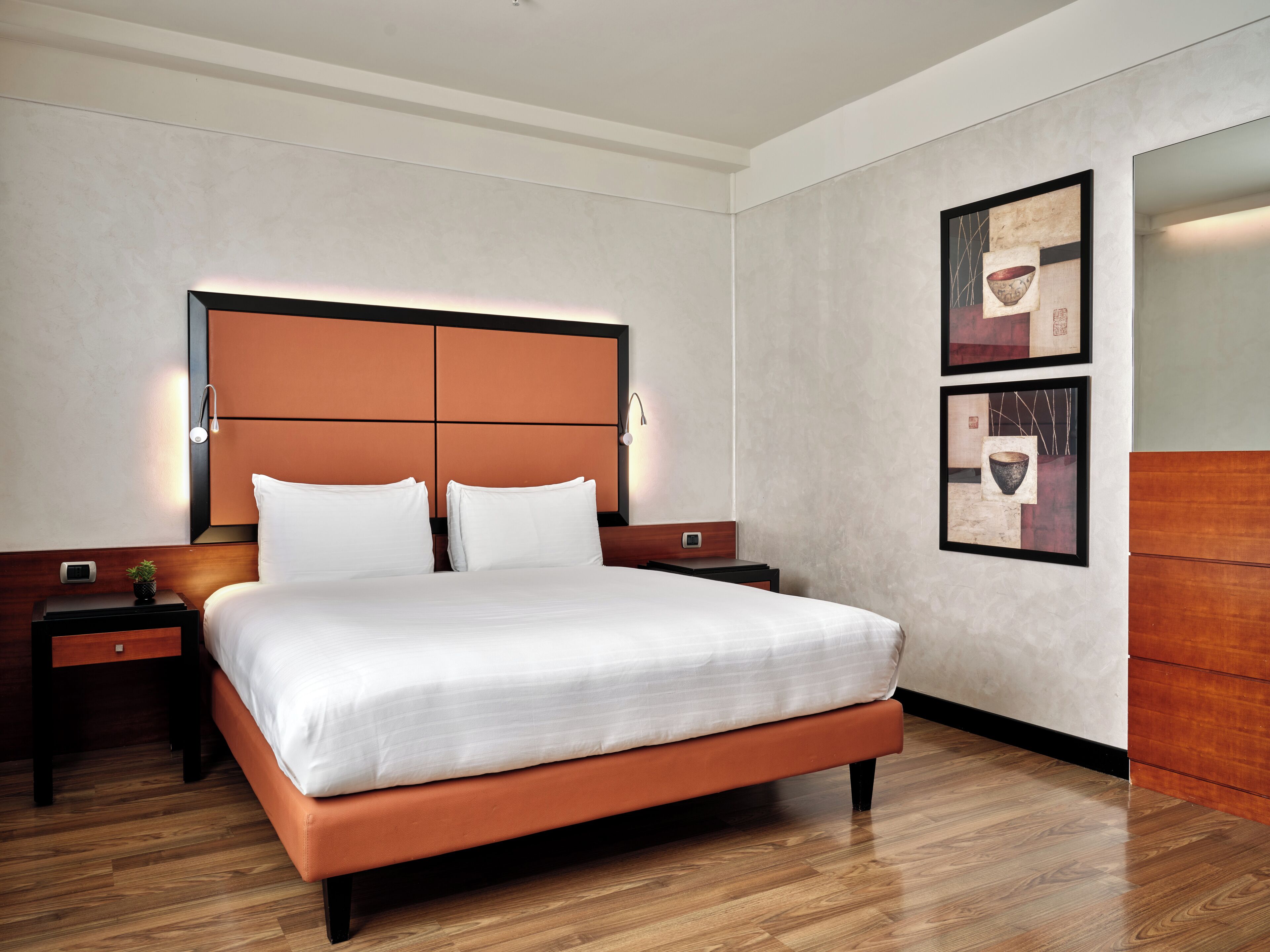 A modern hotel room with a large bed, wooden headboard, two bedside tables, and a wall-mounted artwork.