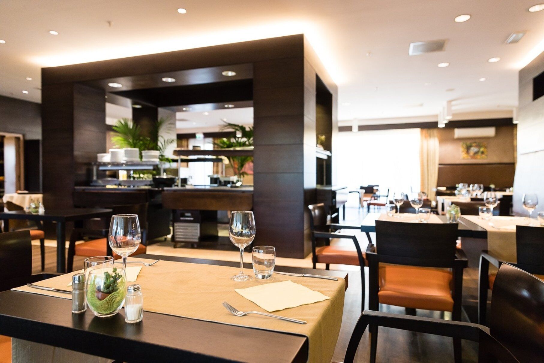 Interno di un ristorante con tavoli apparecchiati per la cena, un'area bar sullo sfondo e vista sulla cucina.