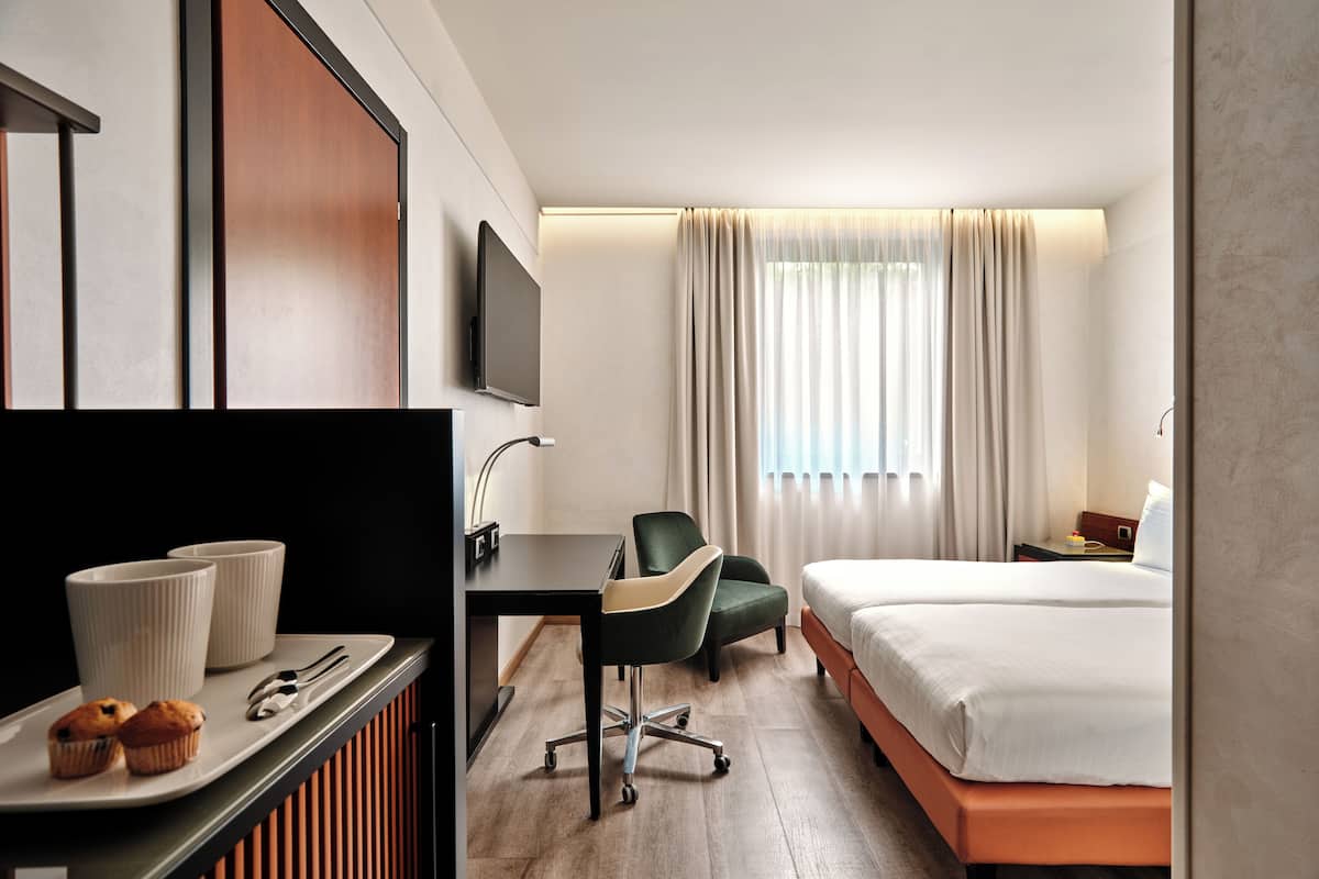 A hotel room with a bed, a desk with a chair, a television, and a window with curtains.