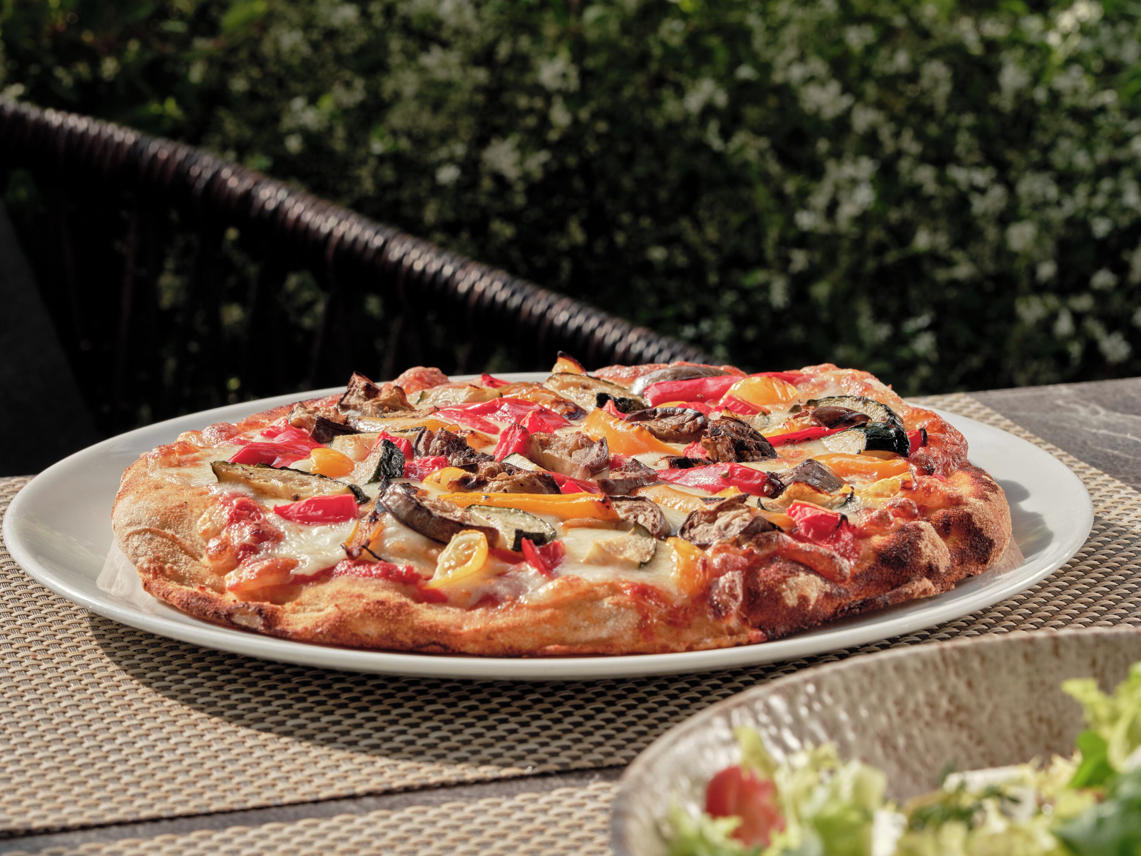 Une pizza garnie de divers ingrédients, servie sur une assiette blanche, posée sur une table recouverte d’une nappe à motifs tissés.