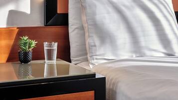 Una cama bien hecha con sábanas blancas, una mesita de noche con un vaso de agua, una pequeña planta en maceta y una lámpara moderna de pared.