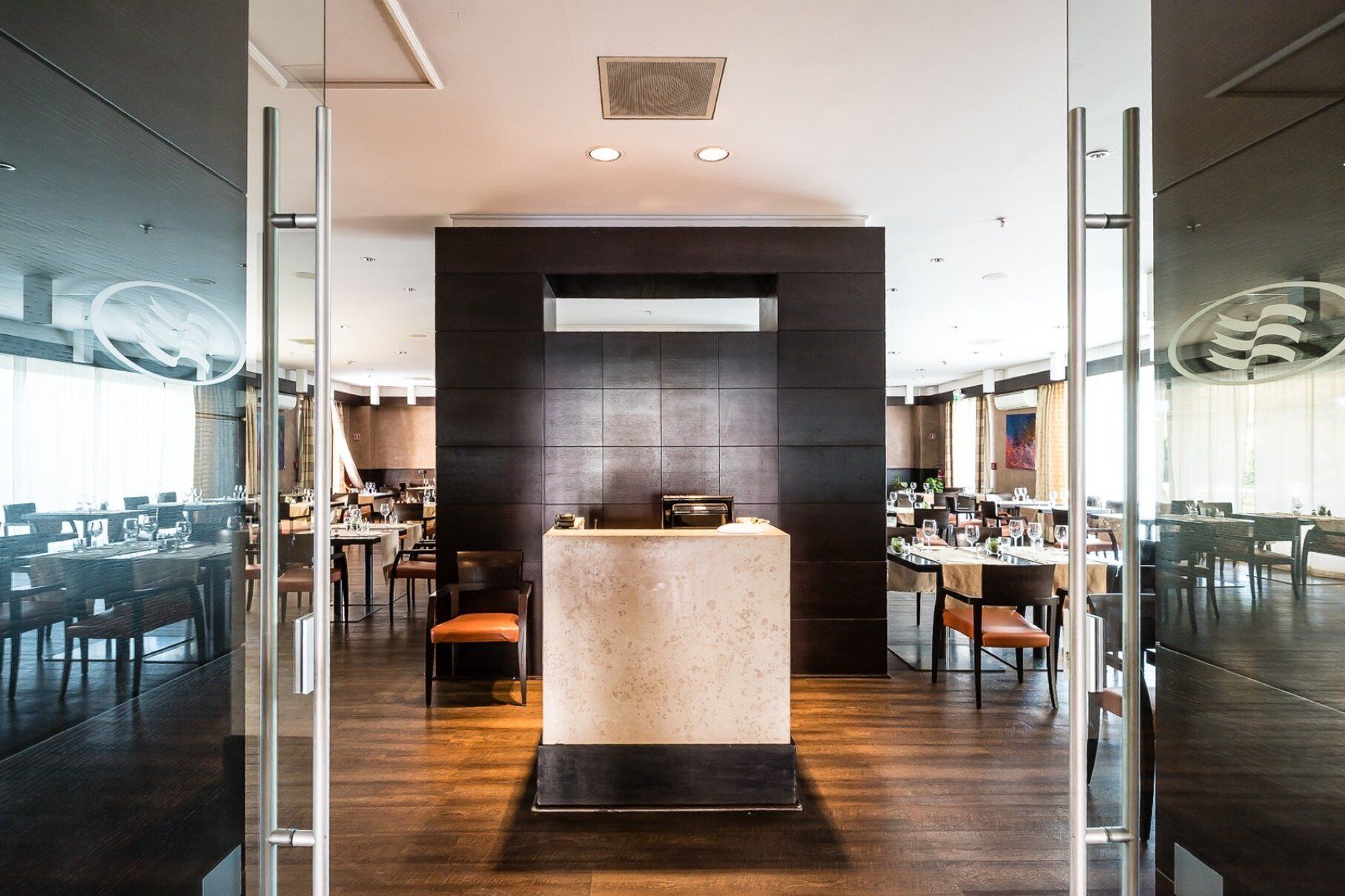 A modern restaurant with a central bar area, multiple dining tables, and a glass partition separating the bar from the dining area.