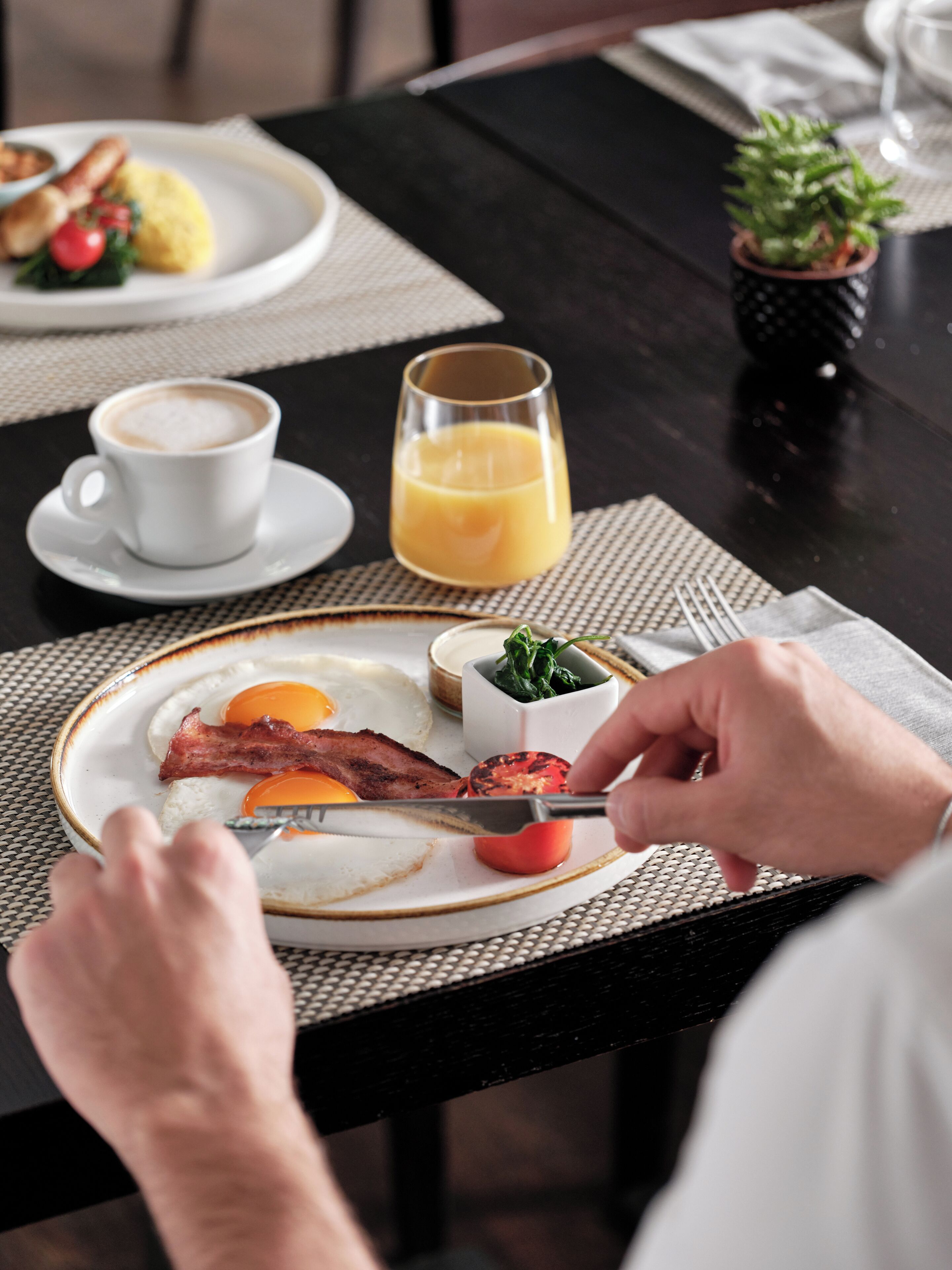 Une table mise pour le petit-déjeuner avec une assiette contenant des œufs, du bacon et une tomate, une tasse de café et un verre de jus d’orange.