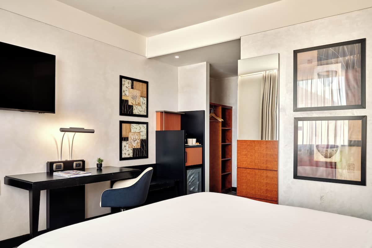 A modern hotel room with a large bed, a desk with a chair, a flat-screen TV, and artwork on the walls.