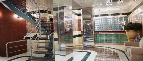 A modern lobby with a marble floor, a staircase, a reception desk, and a large potted plant.